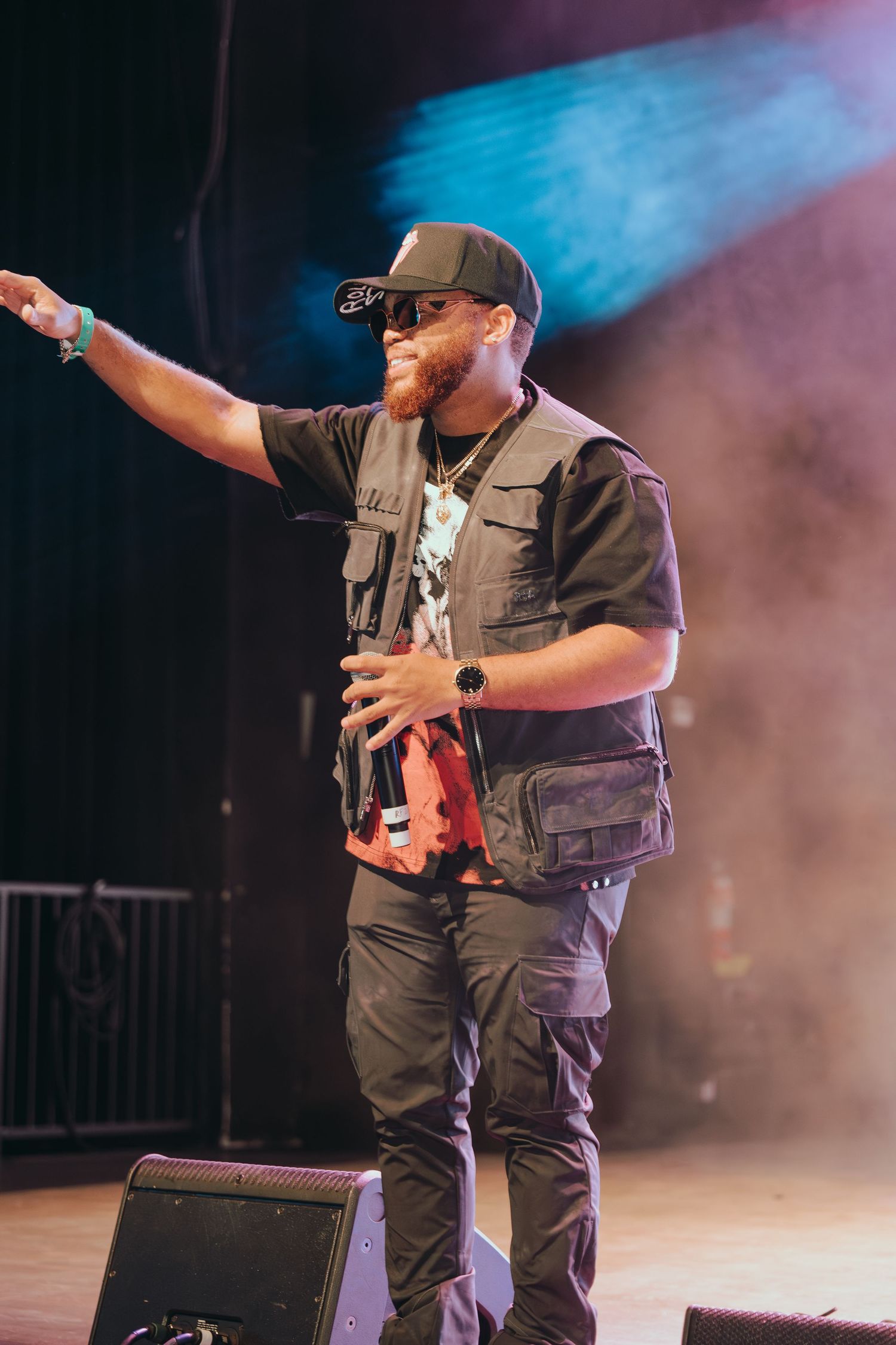 A bearded male performer smiles on stage wearing a black cap and tactical vest at a concert.
