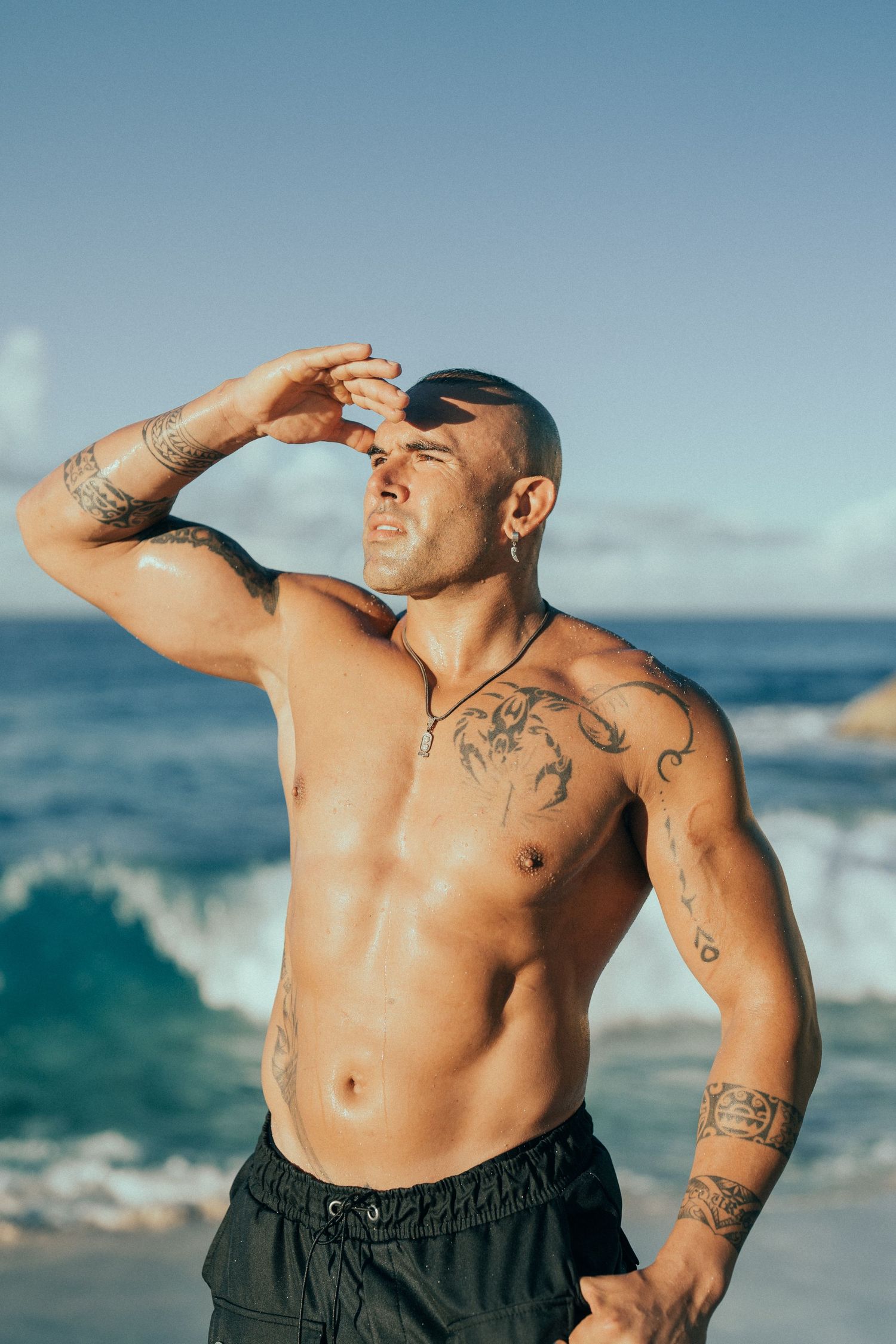 A muscular tattooed shirtless man shields his eyes from the sun while standing on a beach.