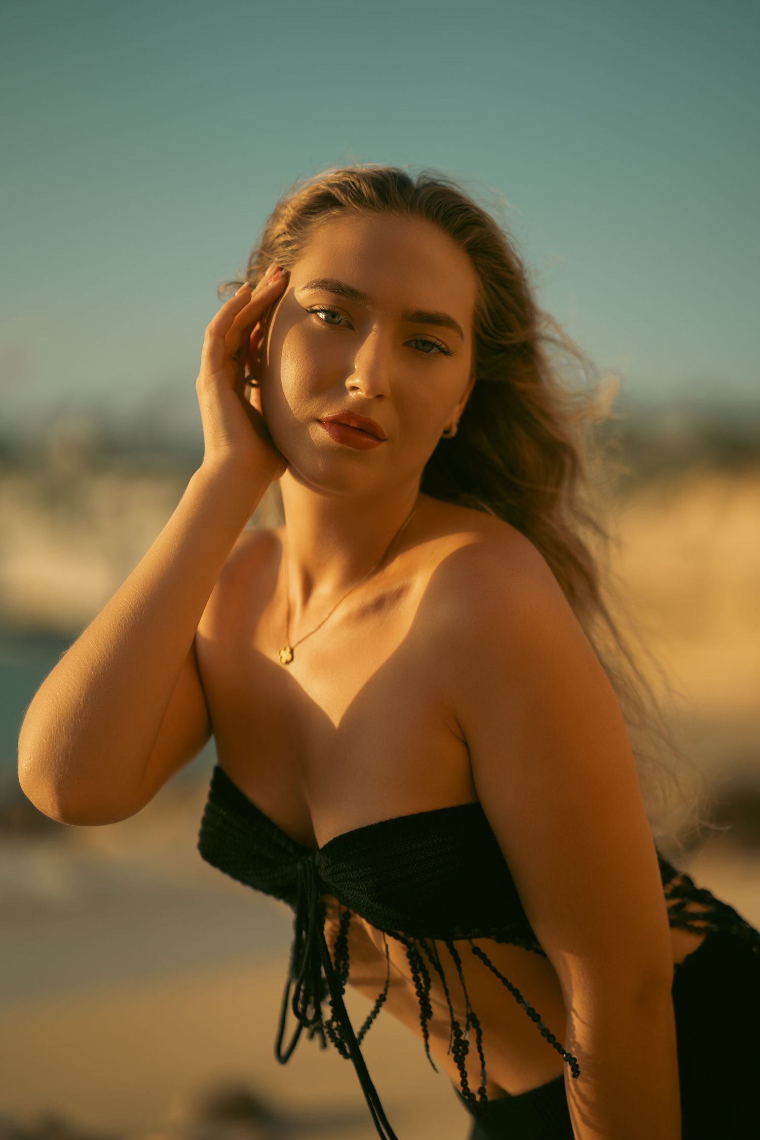 Young woman in black fringed bikini top posing on sandy beach during golden sunset.