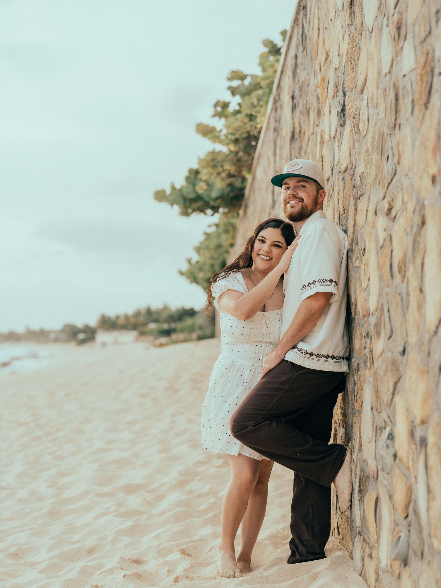 Happy couple leaning against stone wall on sandy beach during engagement photo session.