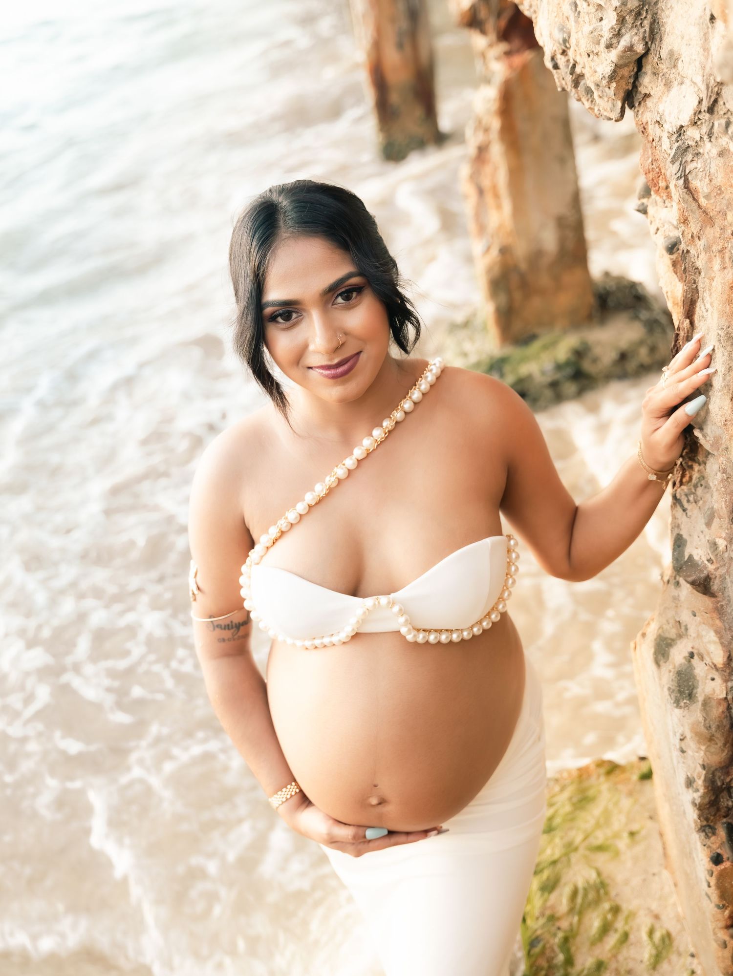 Smiling pregnant woman in pearl bikini top leans against rocky ocean wall at beach.