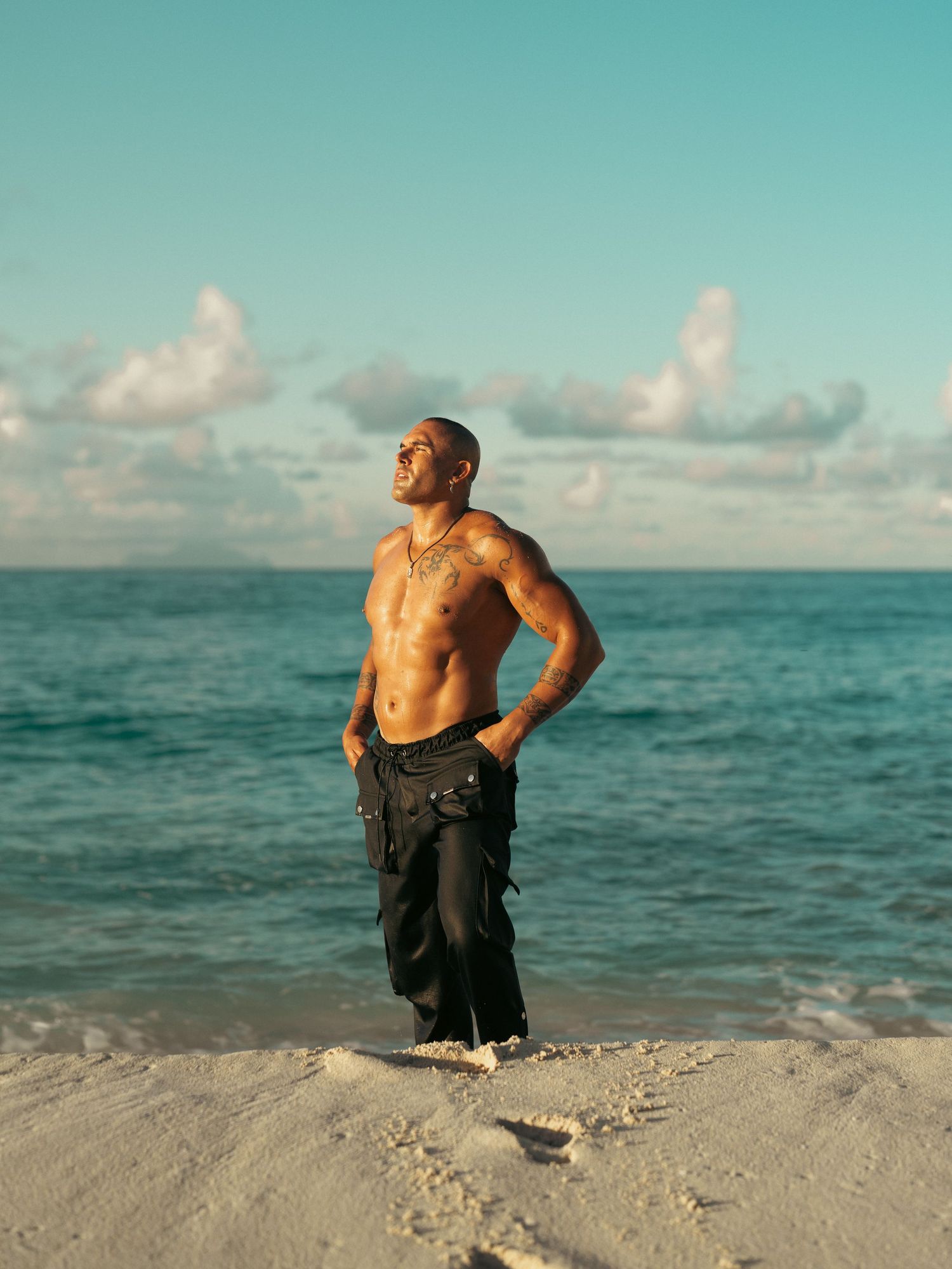 A muscular shirtless man with tattoos stands smiling on a white sand beach under a blue sky.