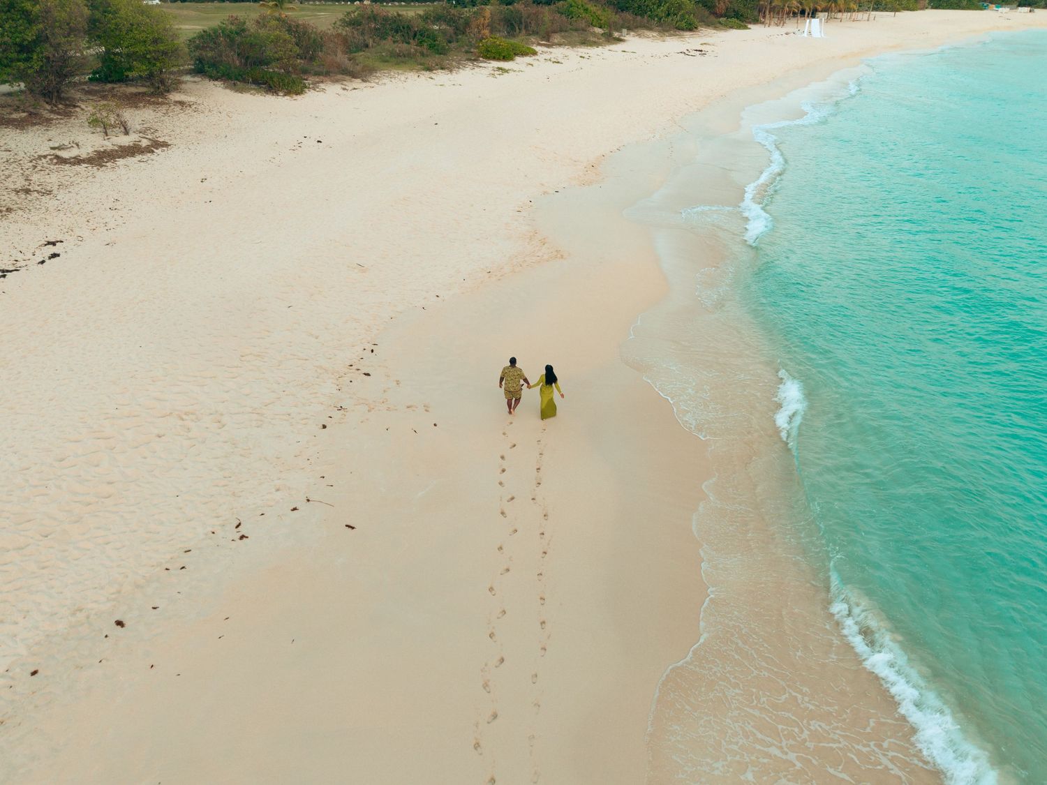 Aerial view of couple holding hands walking on white sand beach by turquoise sea.