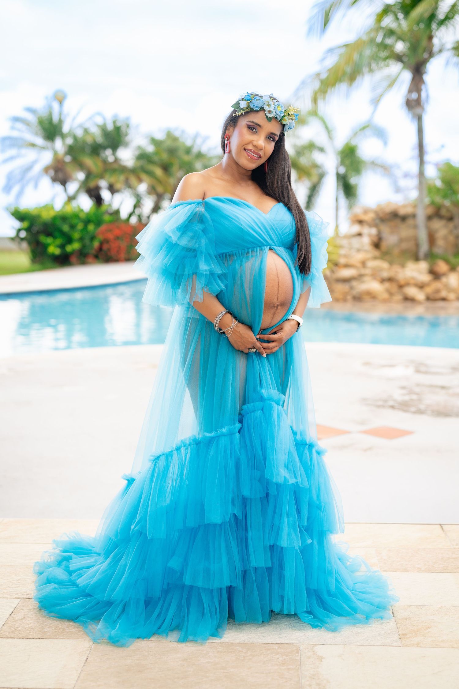 Pregnant woman in elegant blue tulle gown with floral crown poses beside resort pool.
