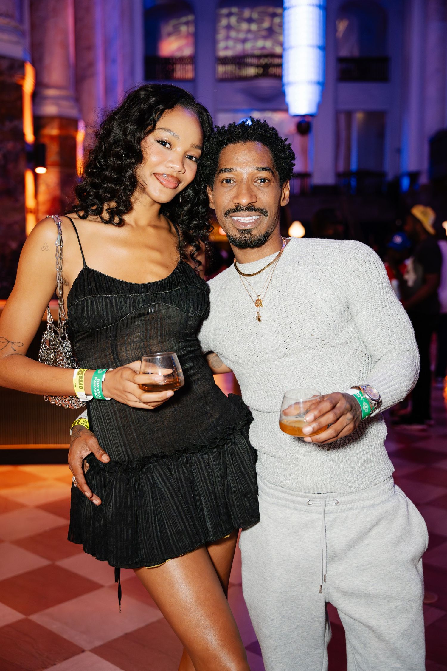 Stylish couple smiles holding drinks at a lively indoor social gathering.