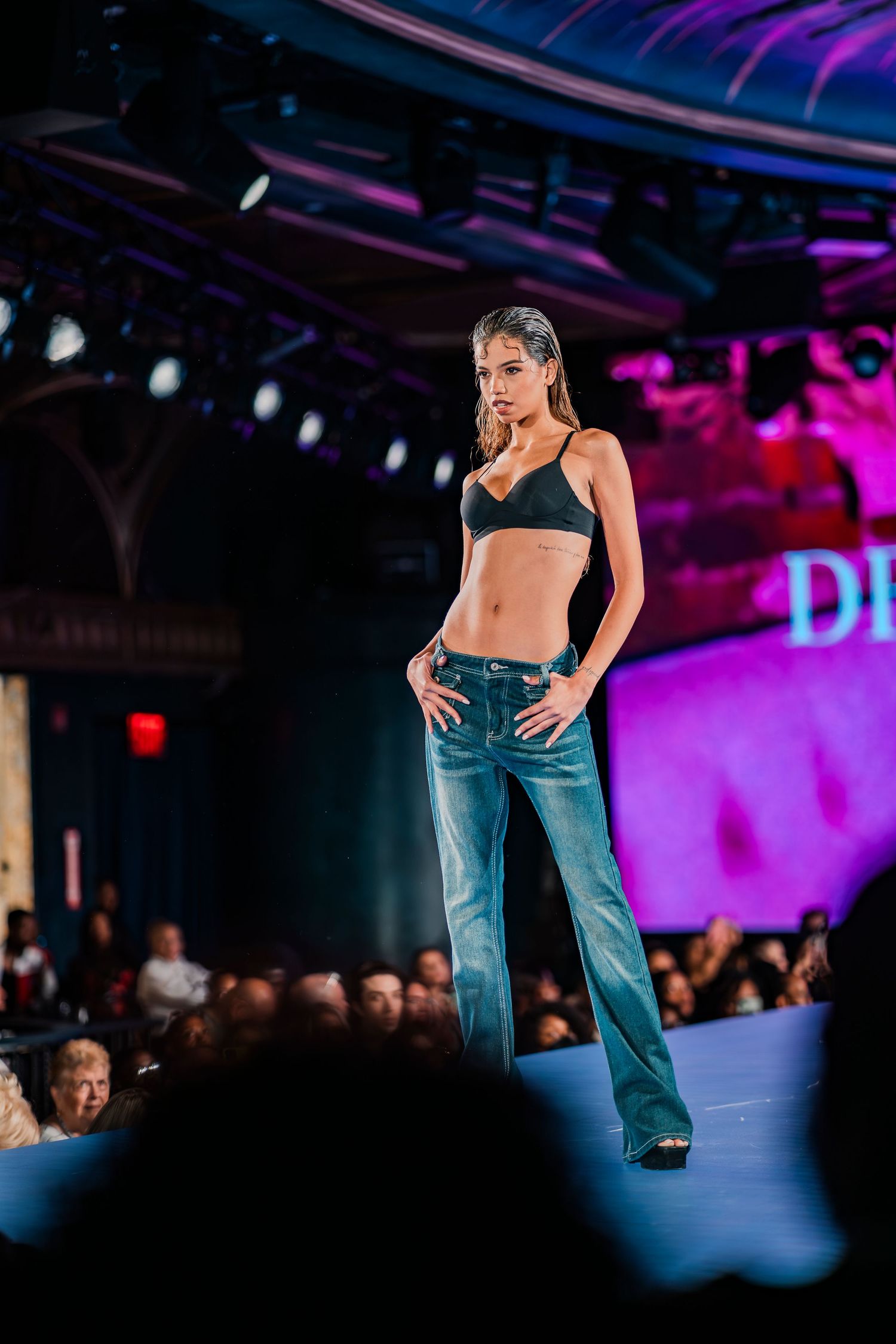 Model walks runway in blue flare jeans and black halter top at a fashion show with colorful stage lighting.