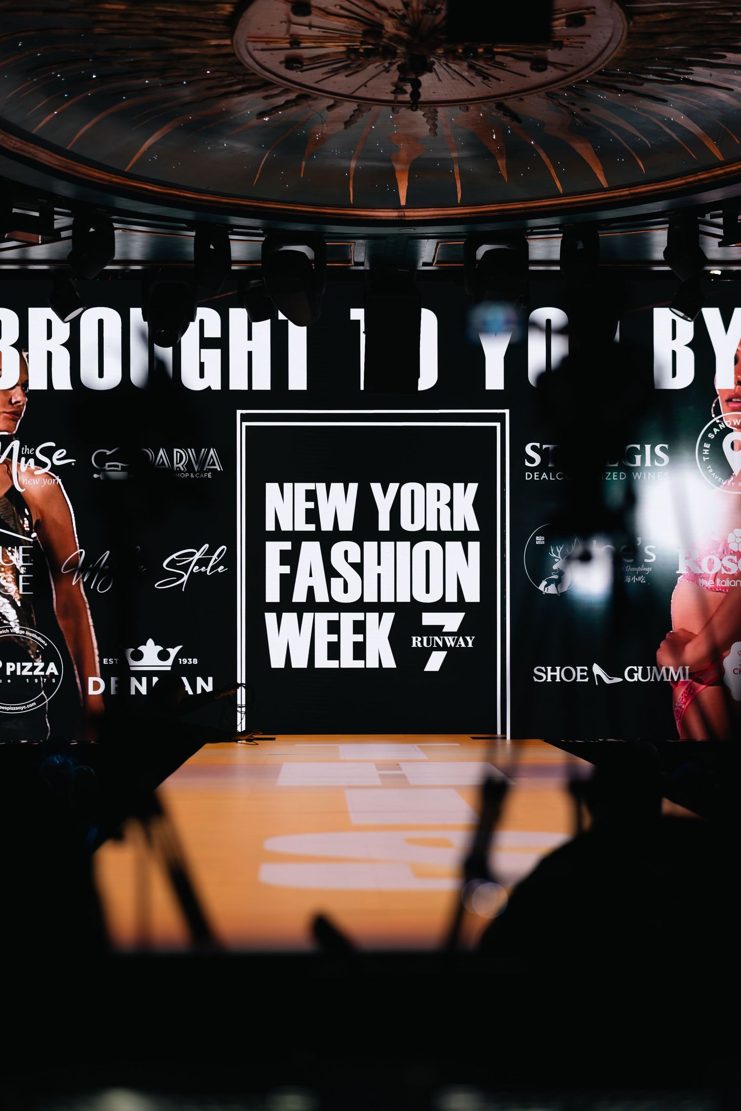 Models walk the runway at New York Fashion Week 7 event on a dramatic stage with sponsor banners and dramatic lighting.