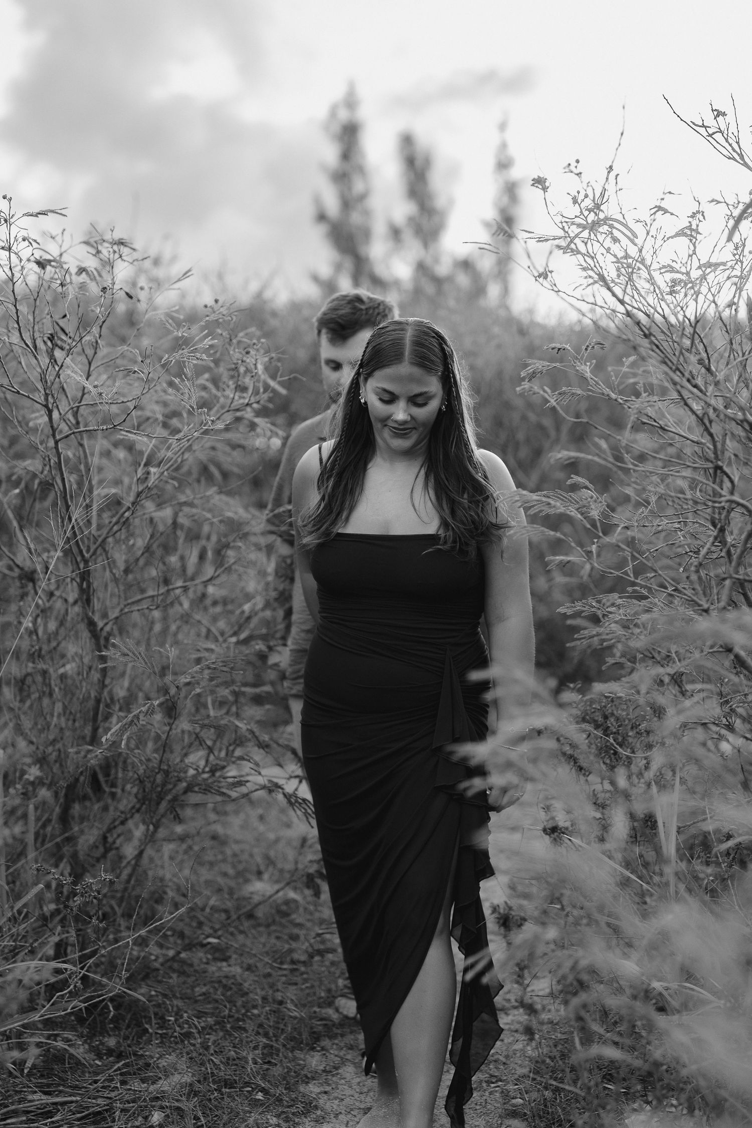 Couple walking through wild brush in black and white engagement photo, woman in elegant black dress.