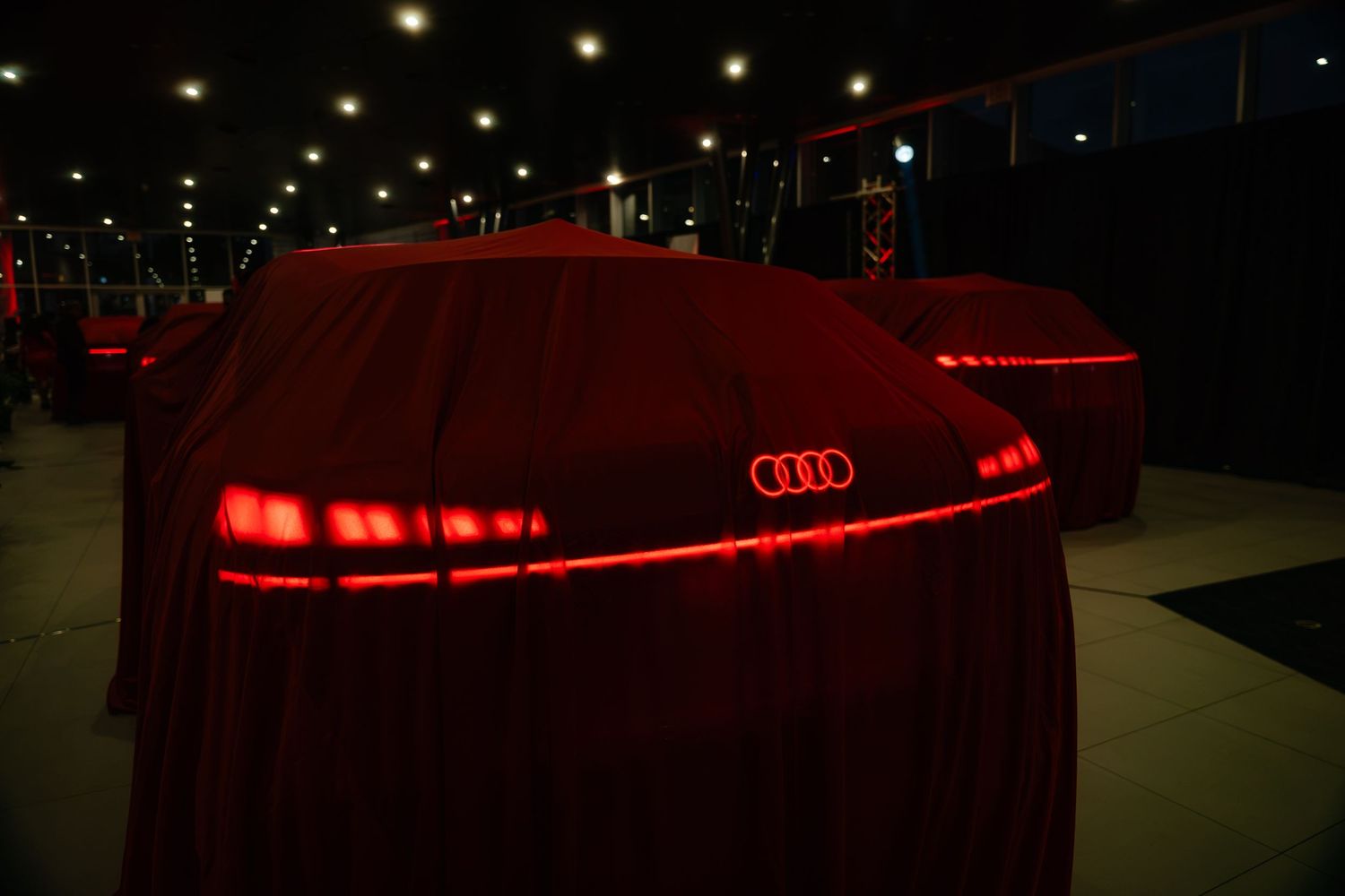 Two Audi cars covered in red velvet sheets at a dramatic car reveal event with glowing red lighting.