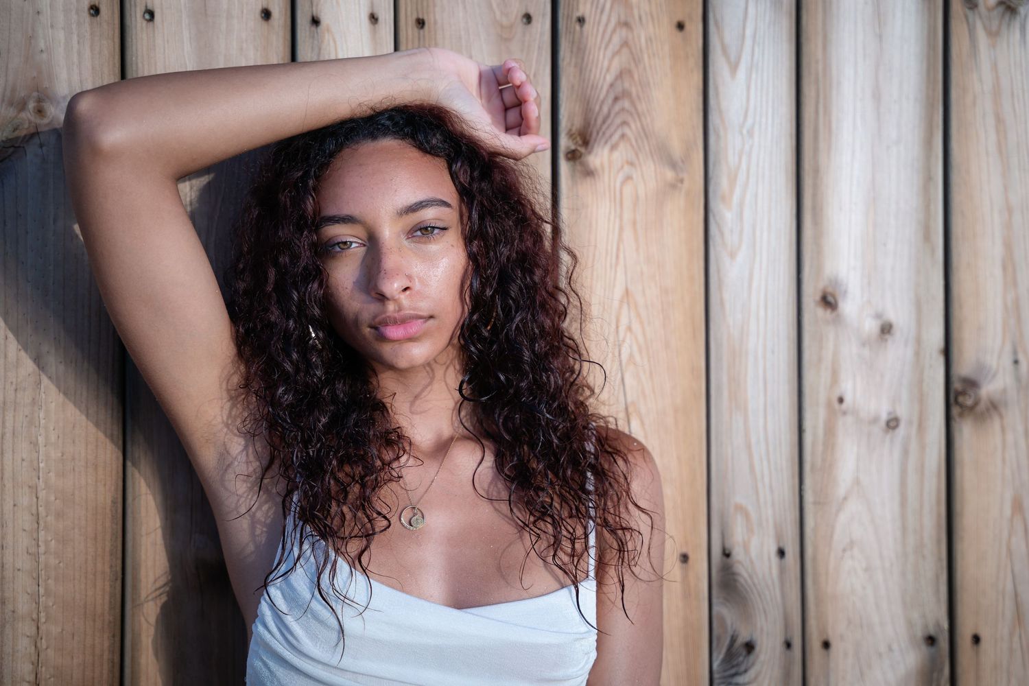 Young woman with curly dark hair in a white top leaning against a wooden plank wall indoors.