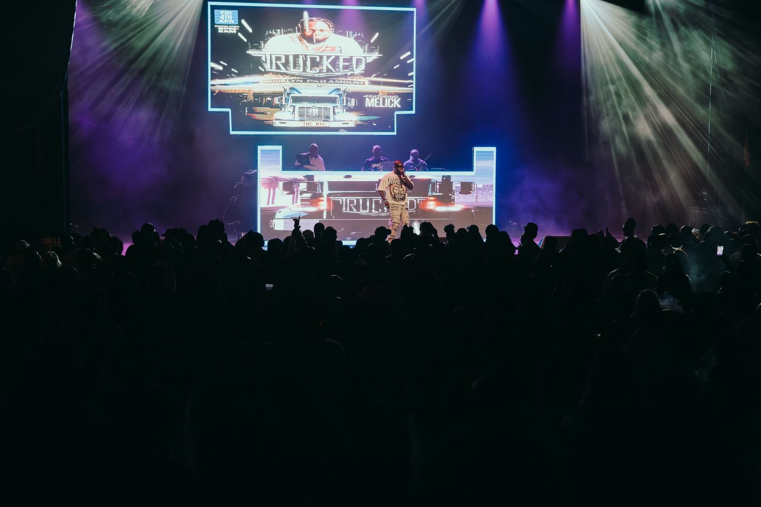 Large crowd watches performers on brightly lit stage at a packed indoor concert venue.