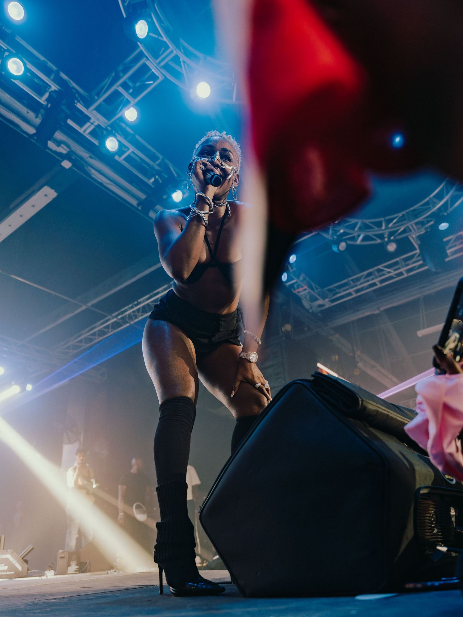 Female performer crouching on stage in black outfit and heeled boots under blue stage lights.