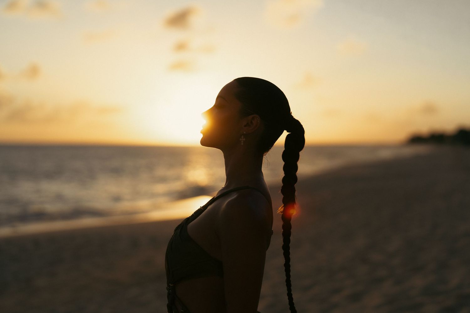 Silhouette of a woman with braided hair standing on a beach during a golden sunset.