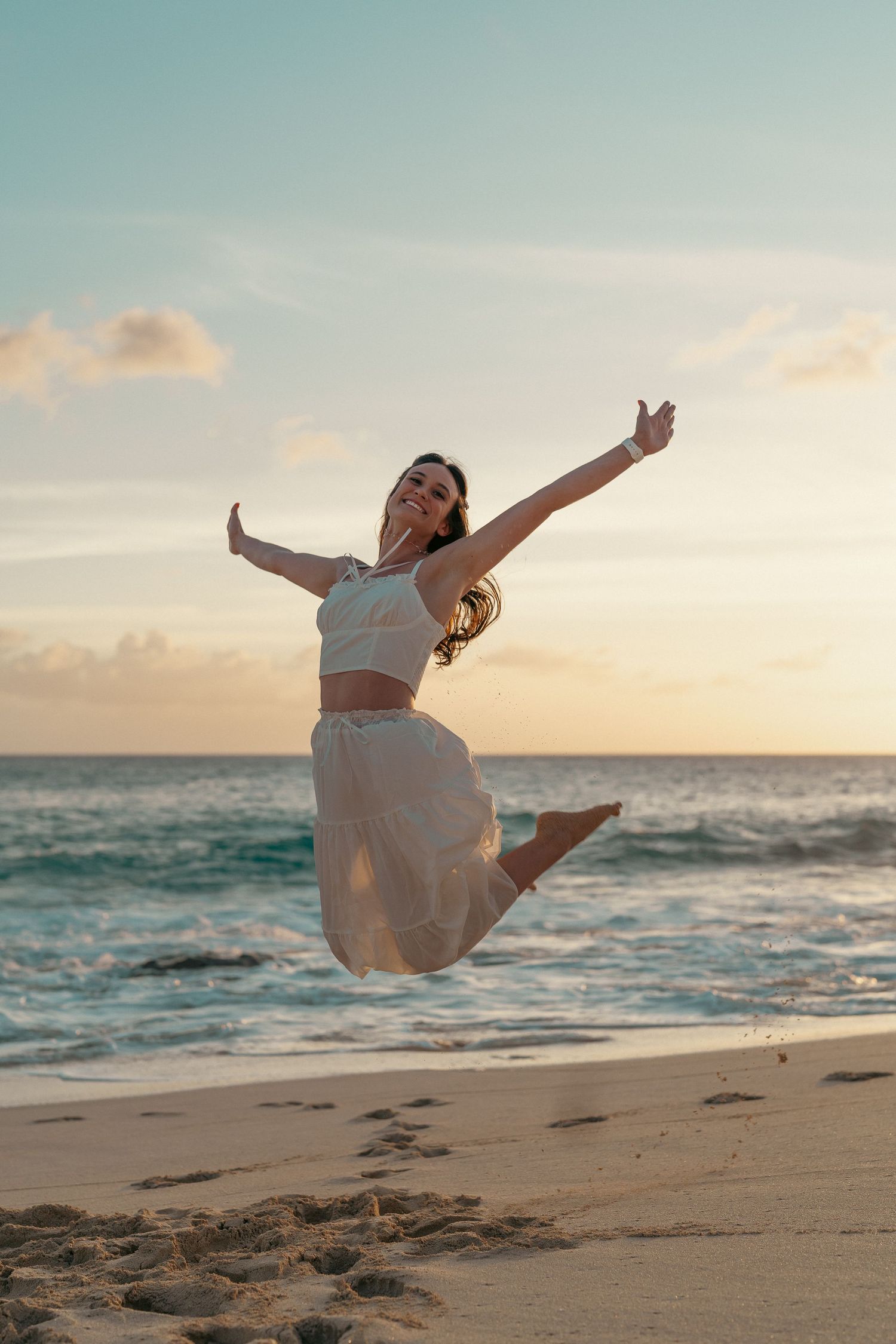Joyful woman in white outfit jumping with arms raised on sandy beach at golden sunset.