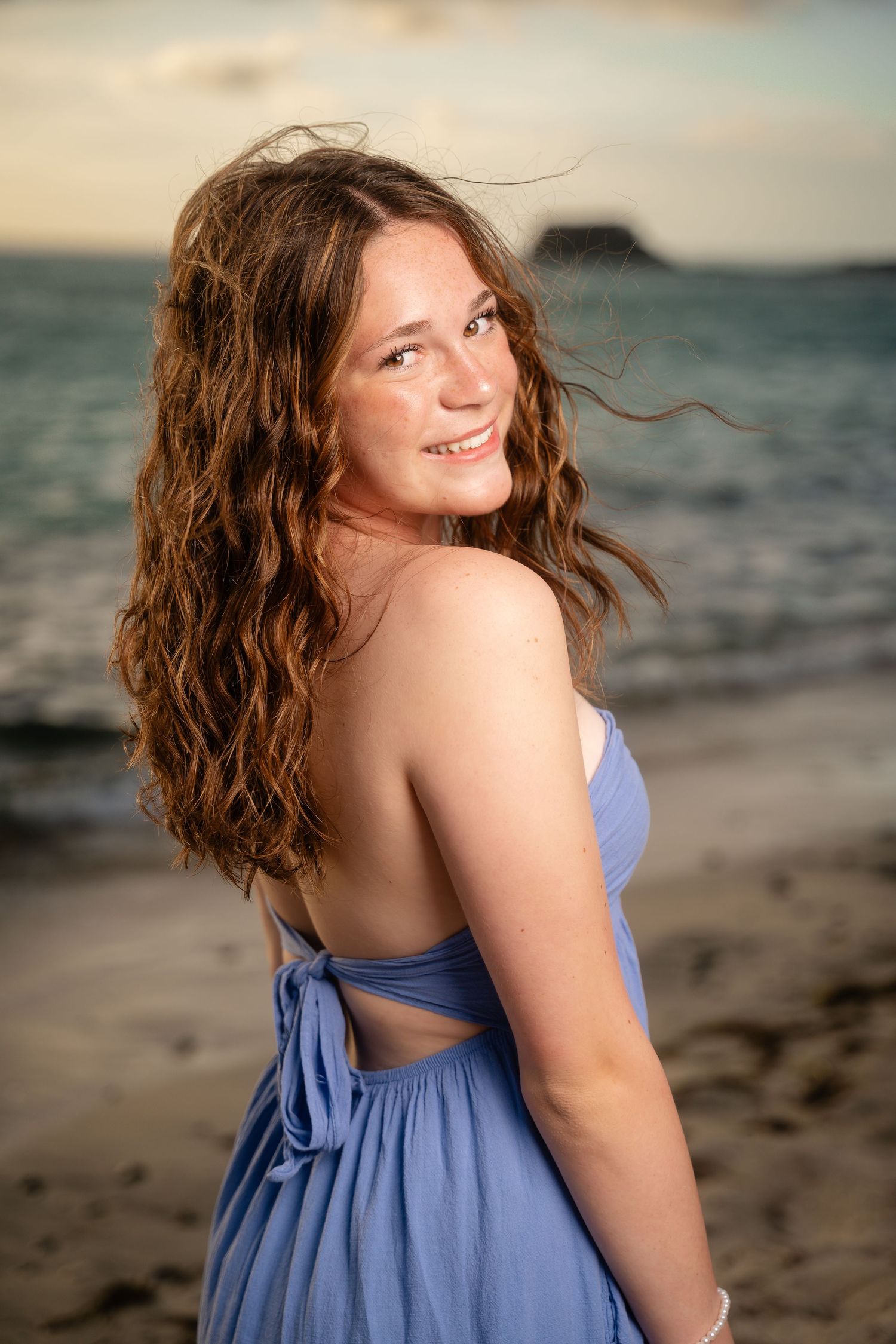 Teenage girl in blue sundress with windswept curly hair smiling over shoulder on beach at sunset.