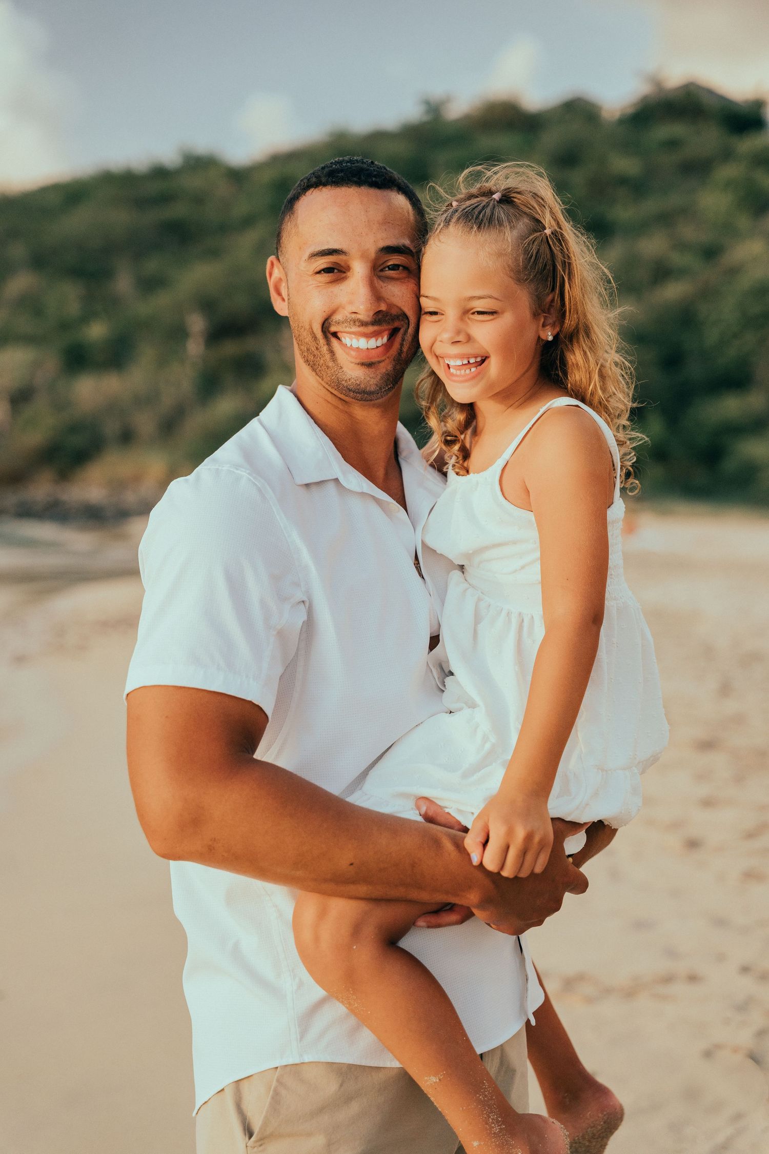 Happy father holding his smiling young daughter in a white dress on a tropical beach at sunset.