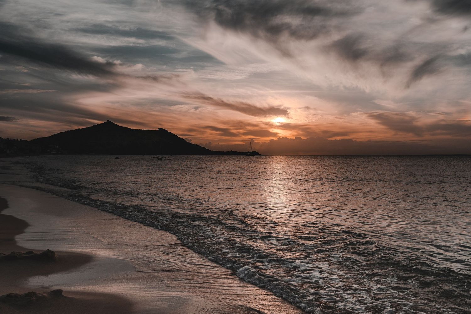 Dramatic sunset over the ocean with a mountain silhouette and moody clouds in the sky.