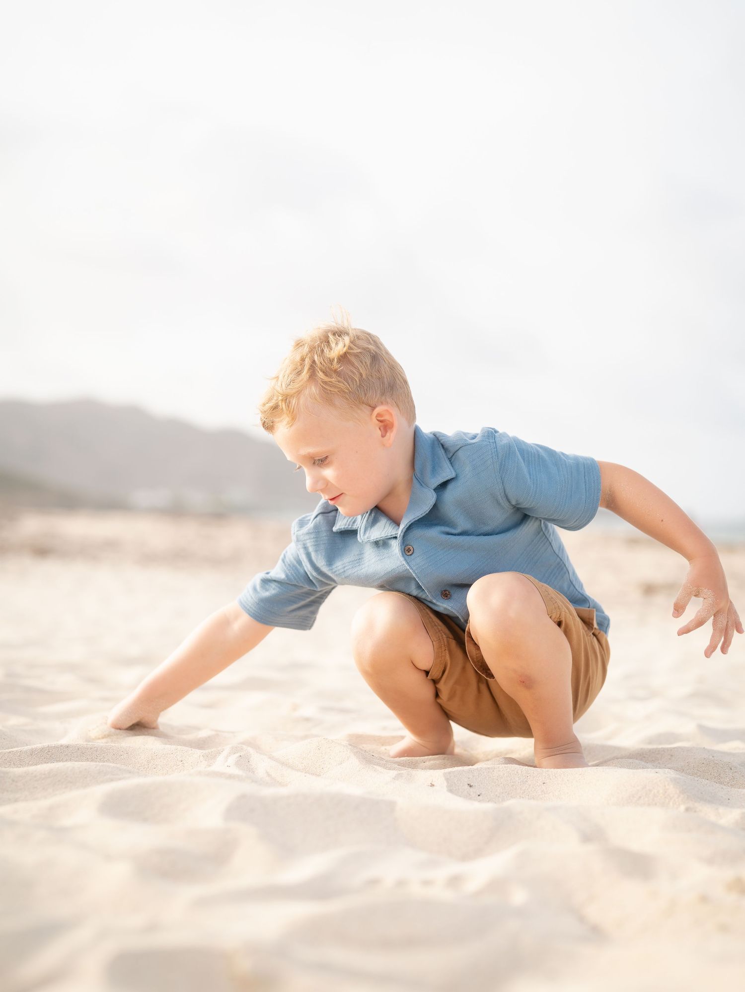 Blonde toddler boy in blue shirt playing in sand at sunny beach.