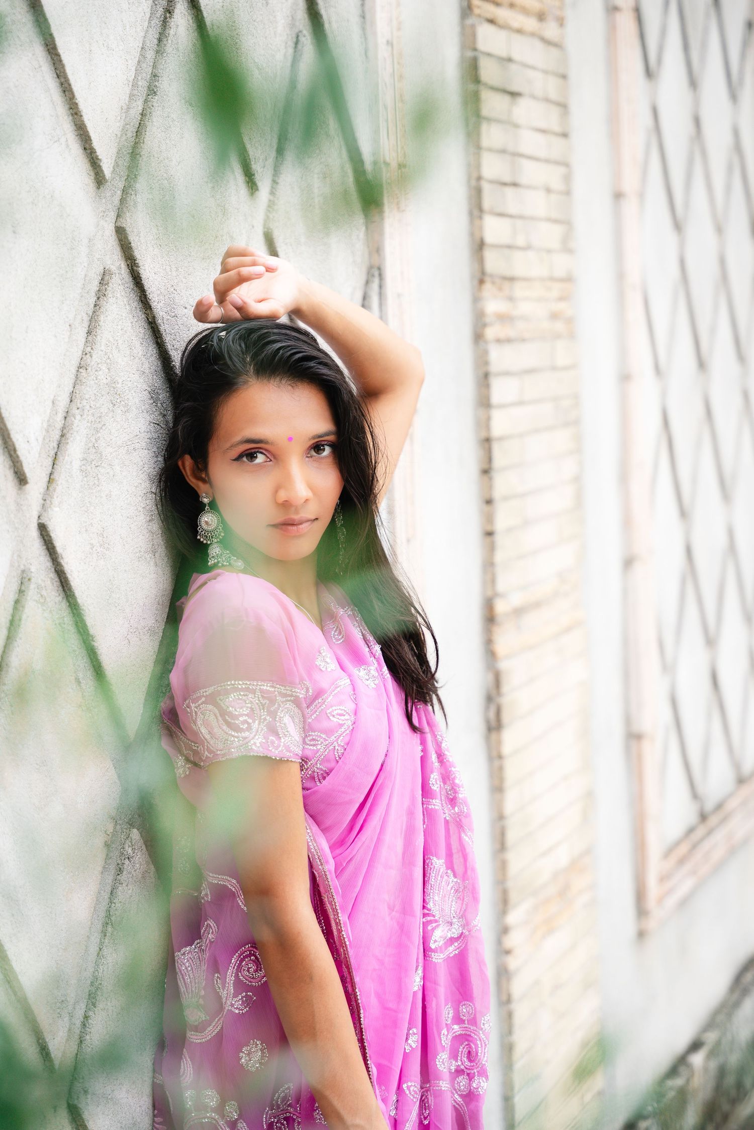 Beautiful woman in pink saree leaning against a weathered wall surrounded by green foliage.