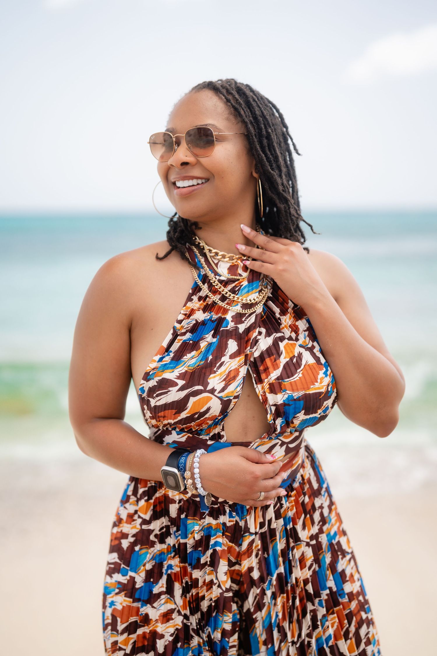 Smiling woman in colorful halter dress and sunglasses poses on a sunny beach with turquoise ocean in background.