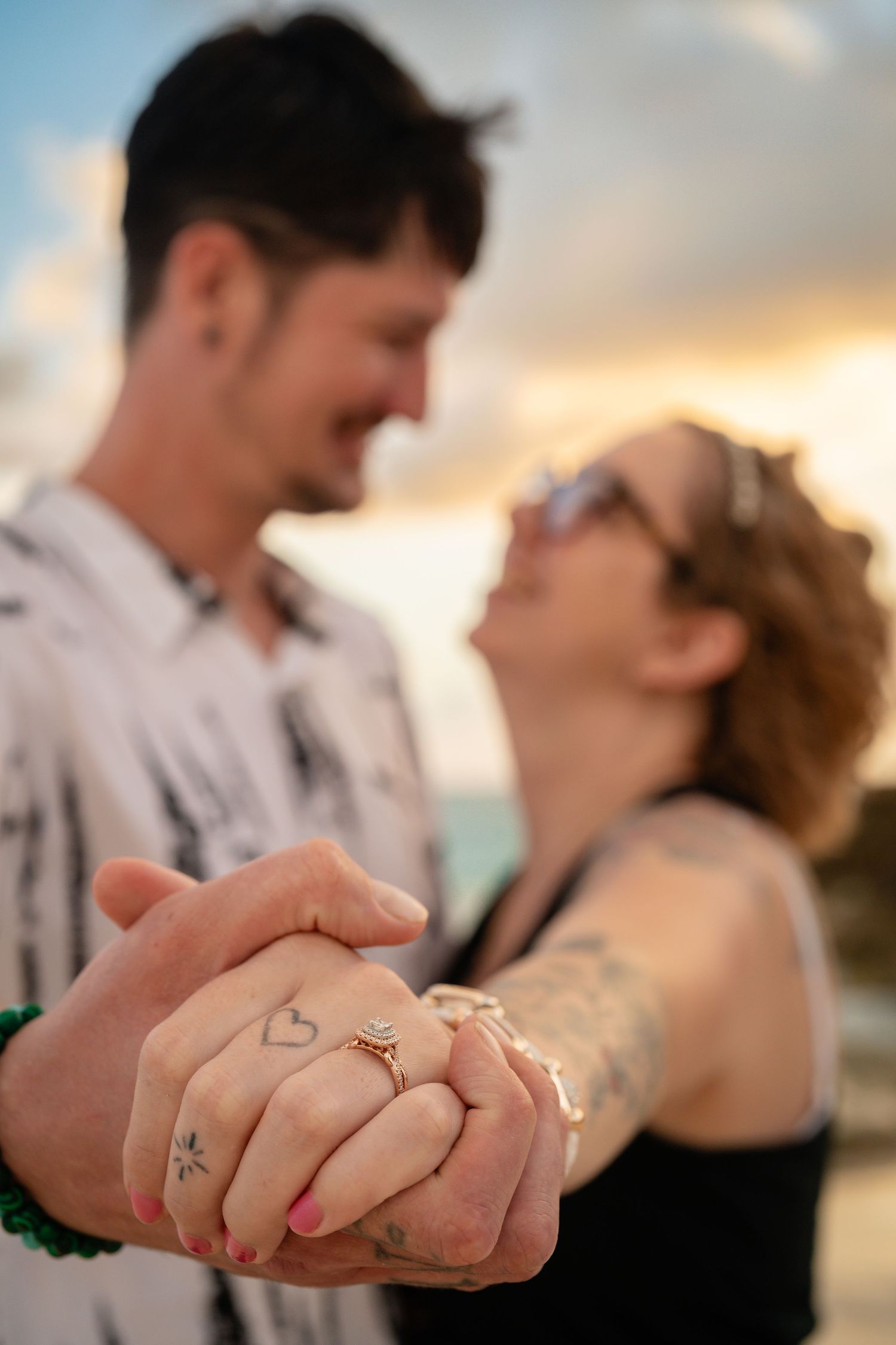 Couple holding hands showing engagement ring with heart tattoos at sunset beach.