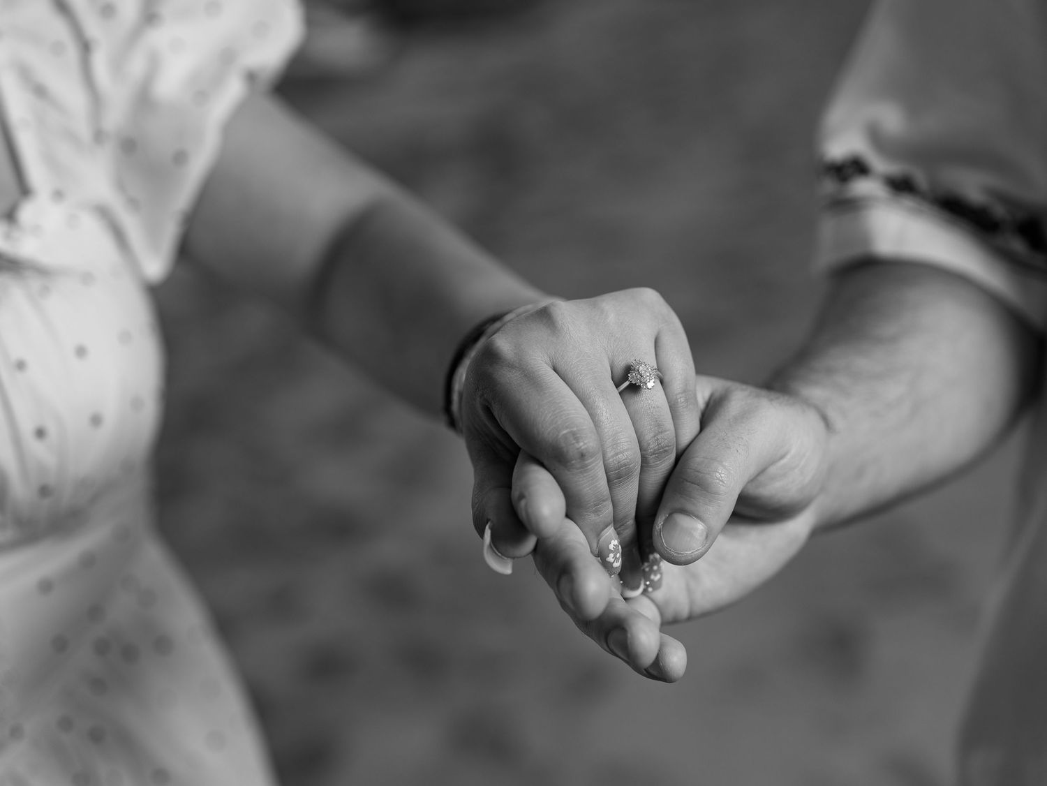 Close-up black and white photo of a couple holding hands showing an engagement ring during a romantic moment.