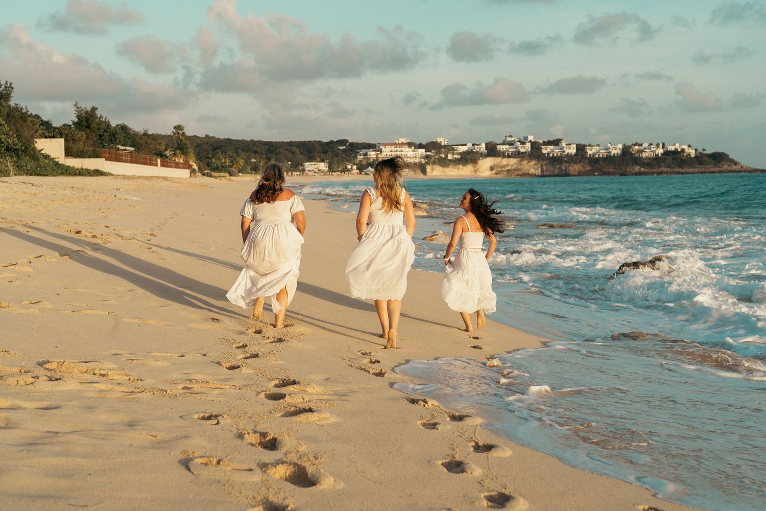 Three girls in white dresses run along a sunny tropical beach near the waves.