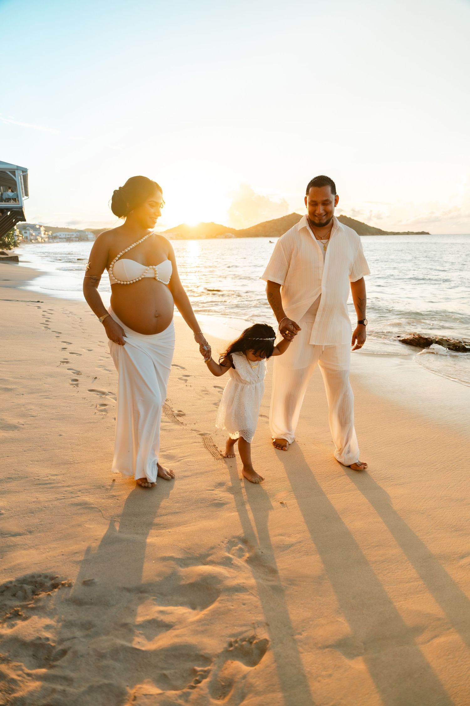 Happy family with pregnant mother in white walks on beach at golden sunset.