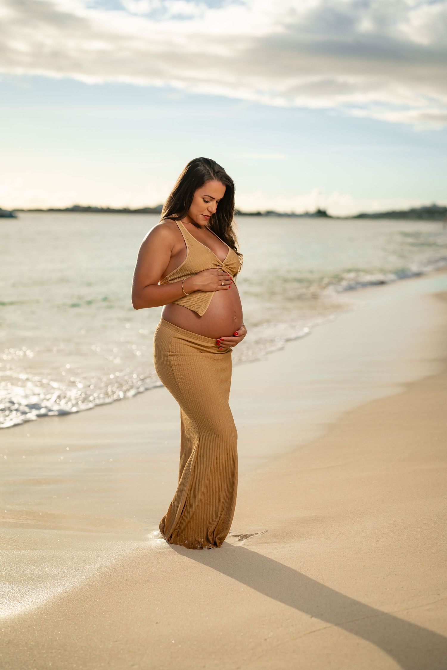 Pregnant woman in gold maternity dress cradling her baby bump on a sandy beach at sunset.