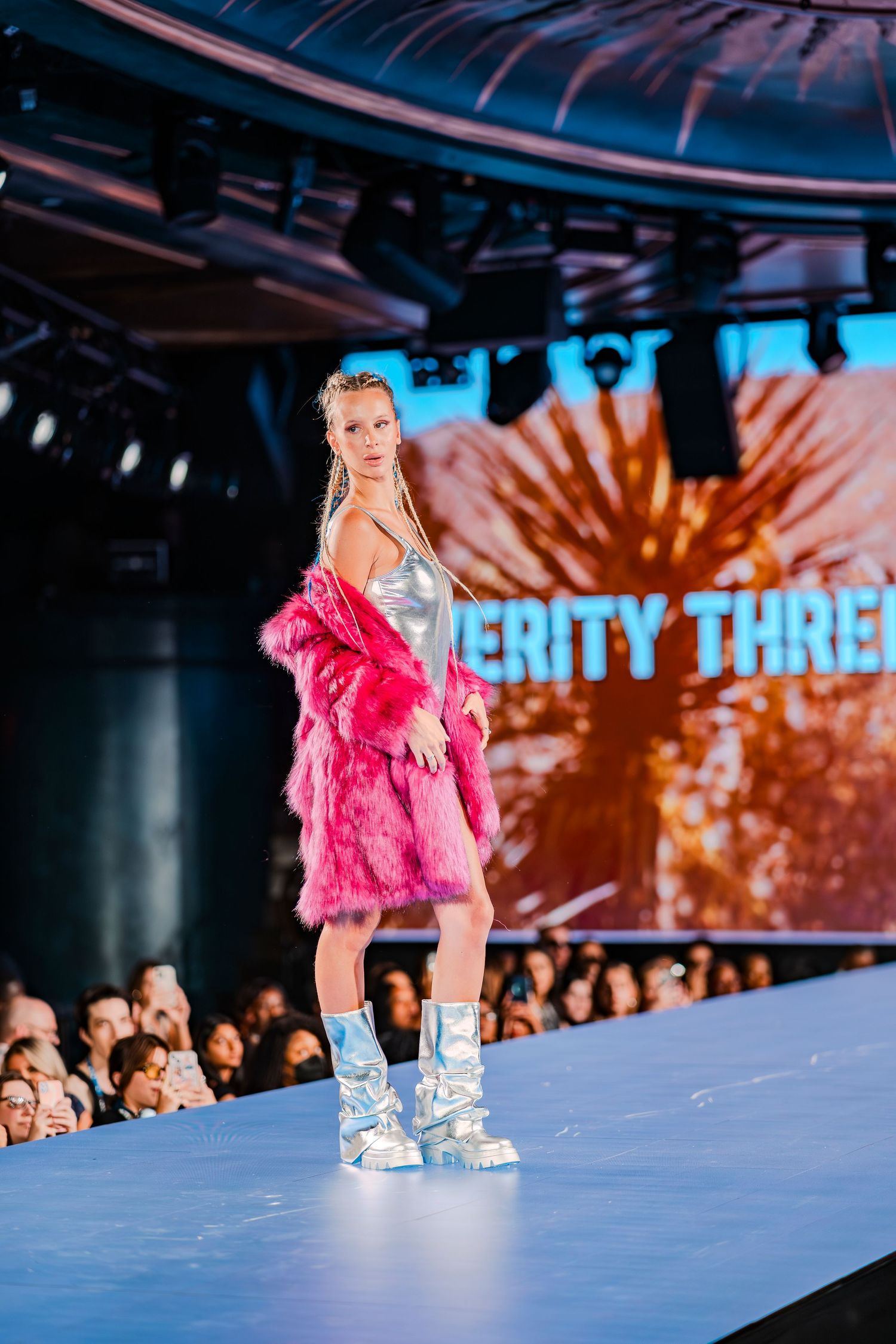 Model walks runway in pink faux fur coat, silver mini dress, and metallic boots at a fashion show.
