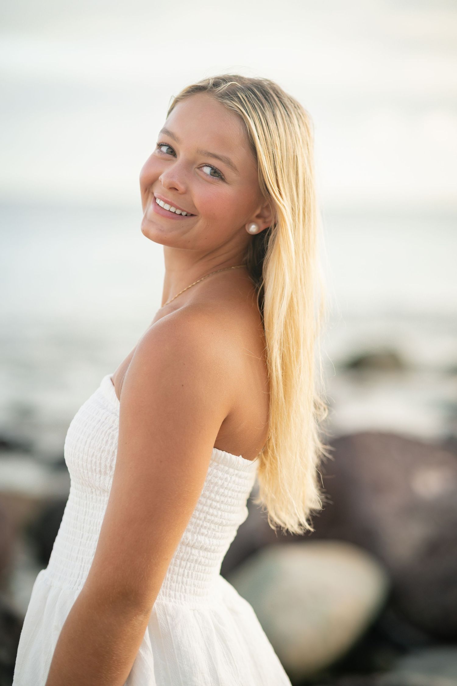 Smiling blonde woman in white strapless dress posing at rocky beach with ocean backdrop.