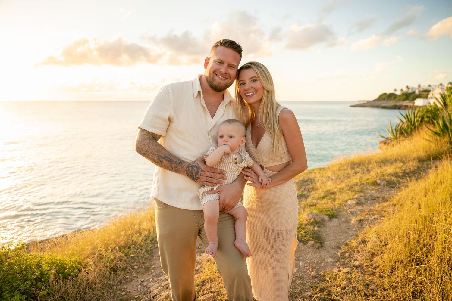 Young family with tattooed dad, mom, and baby posing at golden hour by the ocean on a grassy cliff.