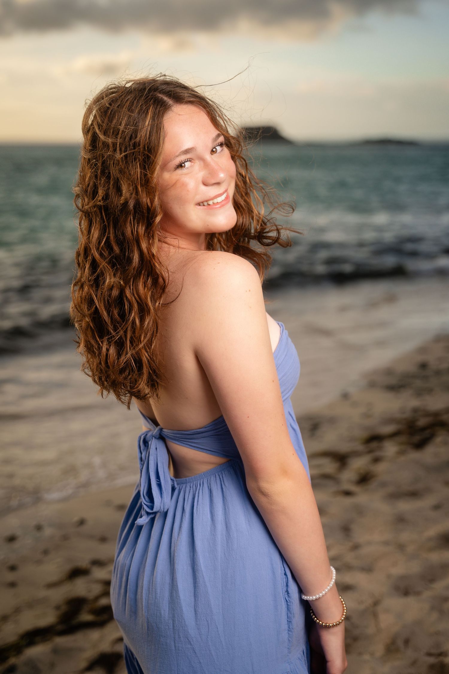 Smiling teenage girl with curly auburn hair in blue backless dress poses on rocky beach at sunset.