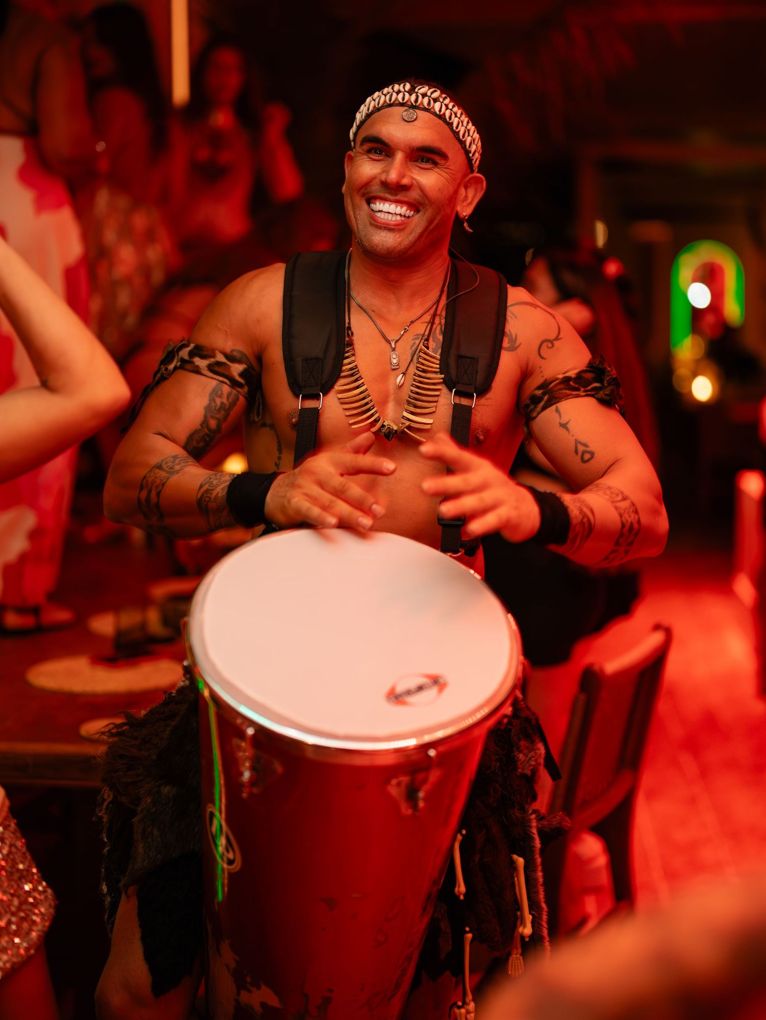 Smiling tattooed male drummer in tribal costume playing red conga drum under red lights.