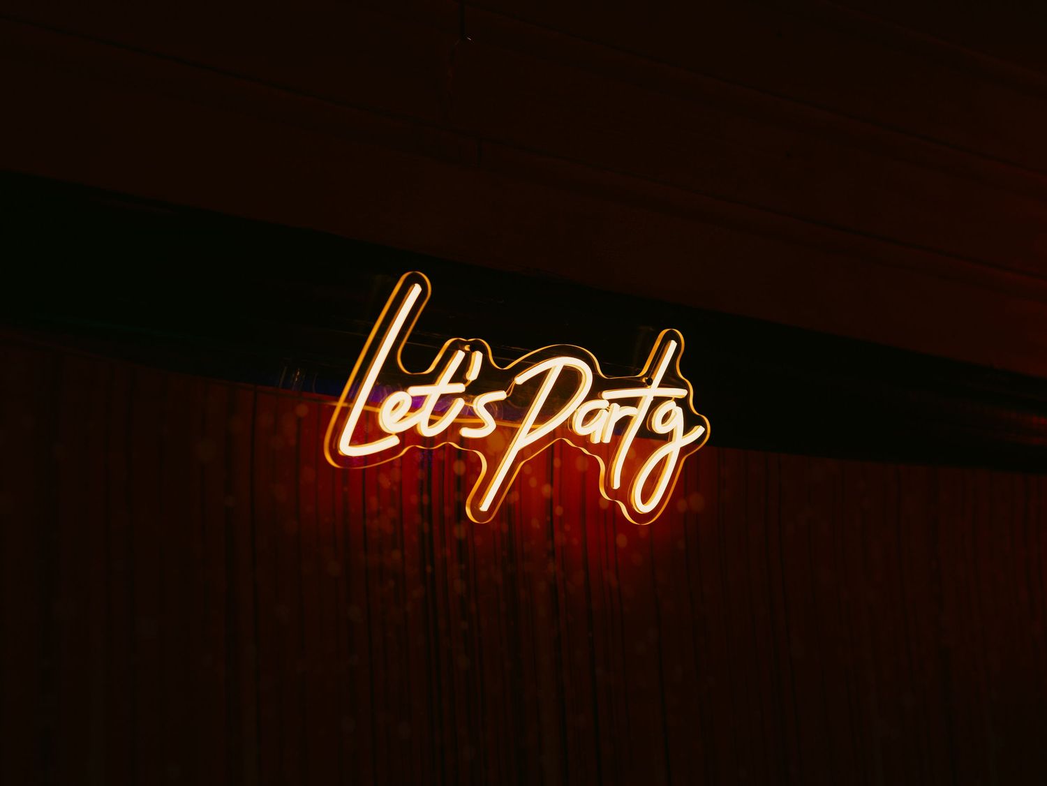 Warm orange neon sign reading Let's Party mounted on a dark wooden wall.