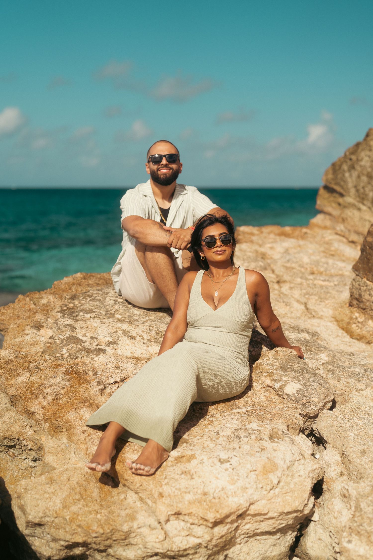 Couple in neutral outfits and sunglasses relaxing on rocky coastal cliffs by turquoise sea.