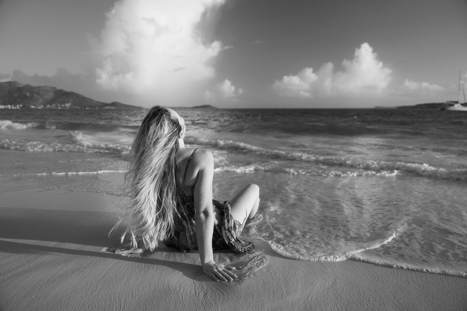 Black and white photo of a long-haired woman sitting on a sandy beach with ocean waves behind her.