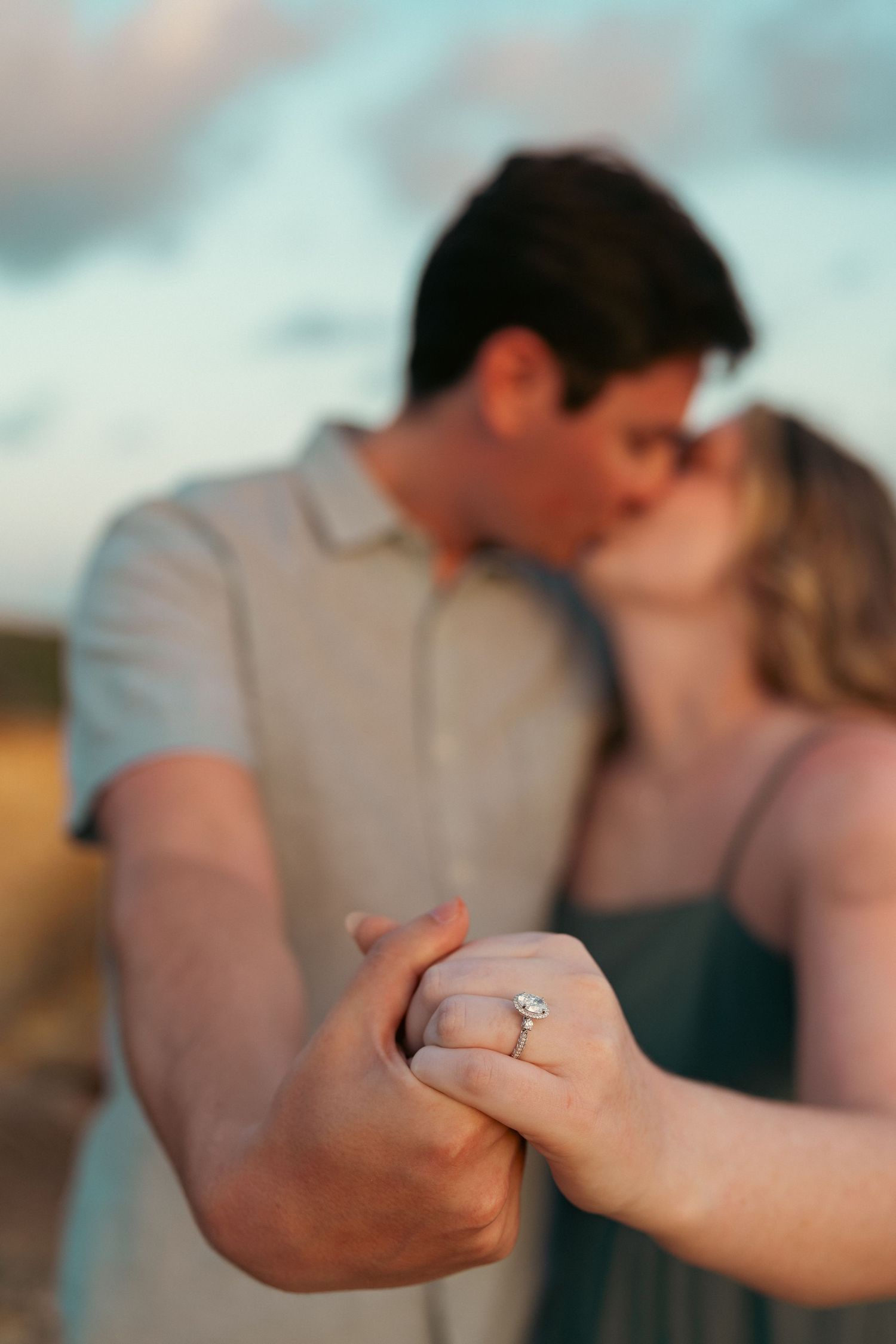 Engaged couple kissing while showcasing diamond engagement ring in golden hour light.