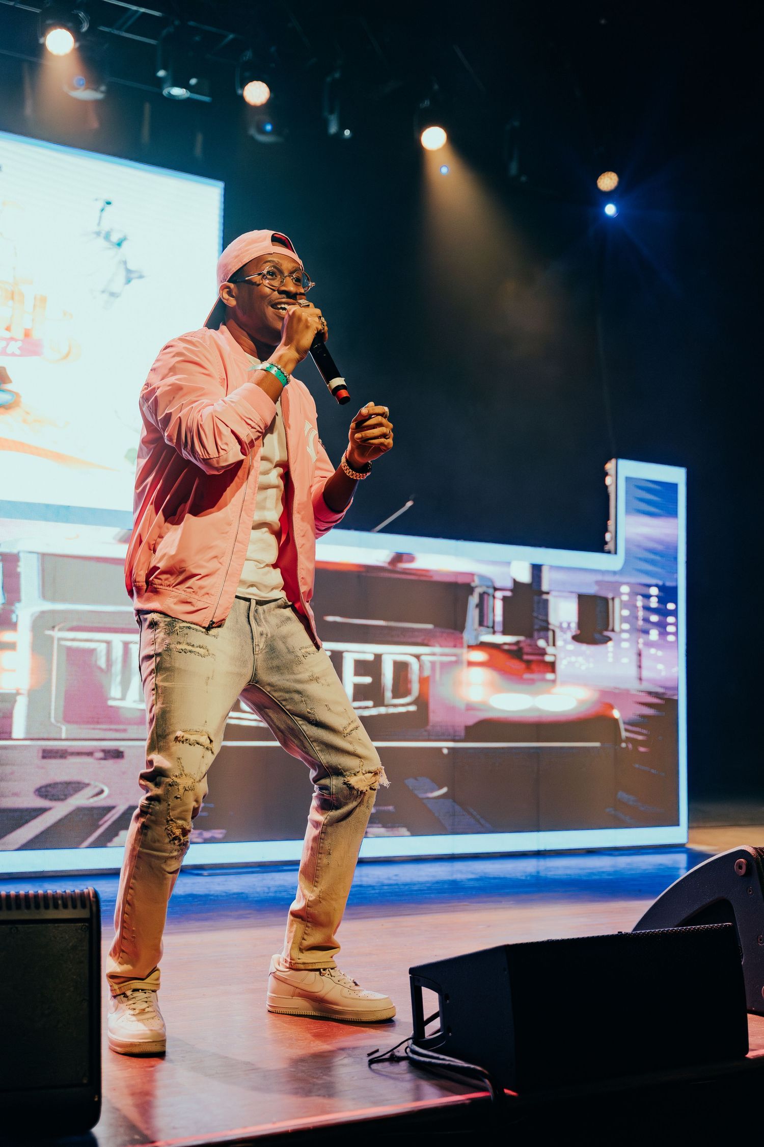 Male performer in pink jacket and cap singing on stage with colorful concert lighting.