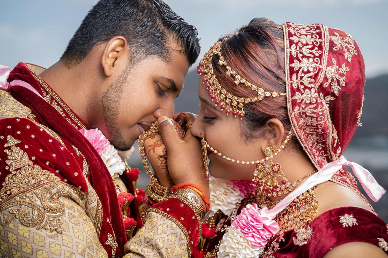 Indian bride and groom in traditional red and gold wedding attire share intimate moment.