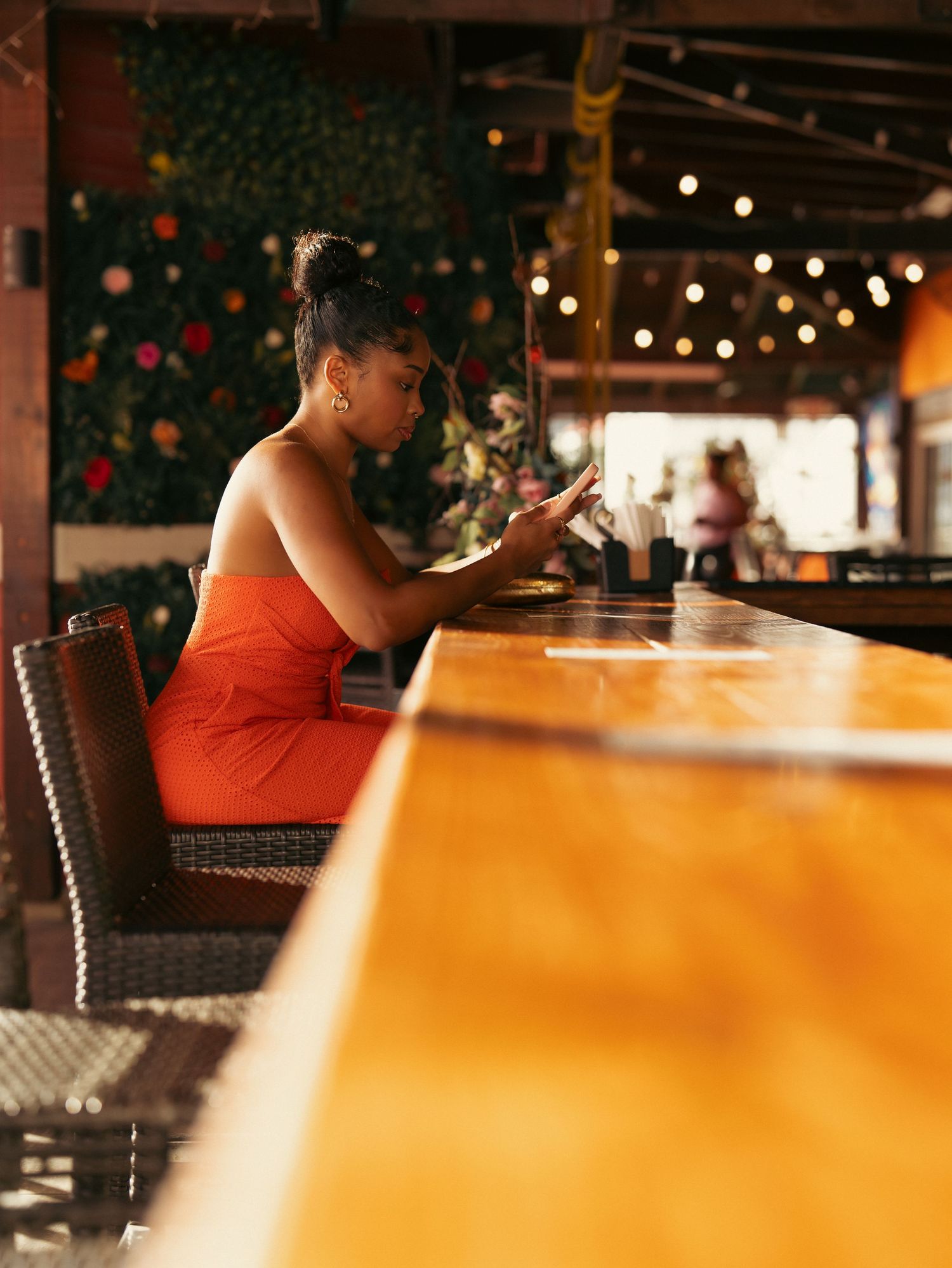 Woman in orange dress sitting at a wooden bar counter looking at her phone in a tropical restaurant.