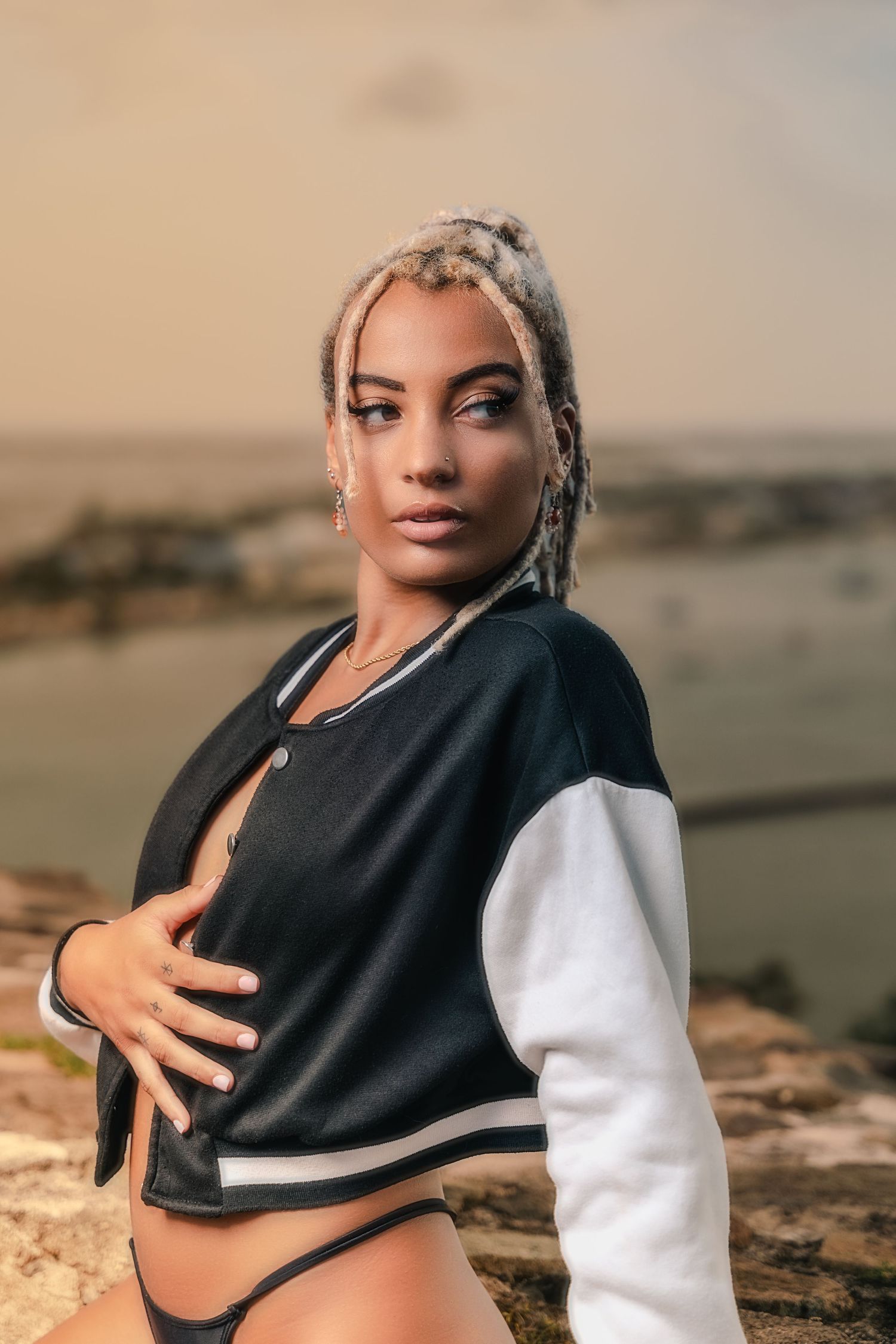 Woman with blonde dreadlocks in a black and white varsity jacket poses on rocky cliff with city view behind her.