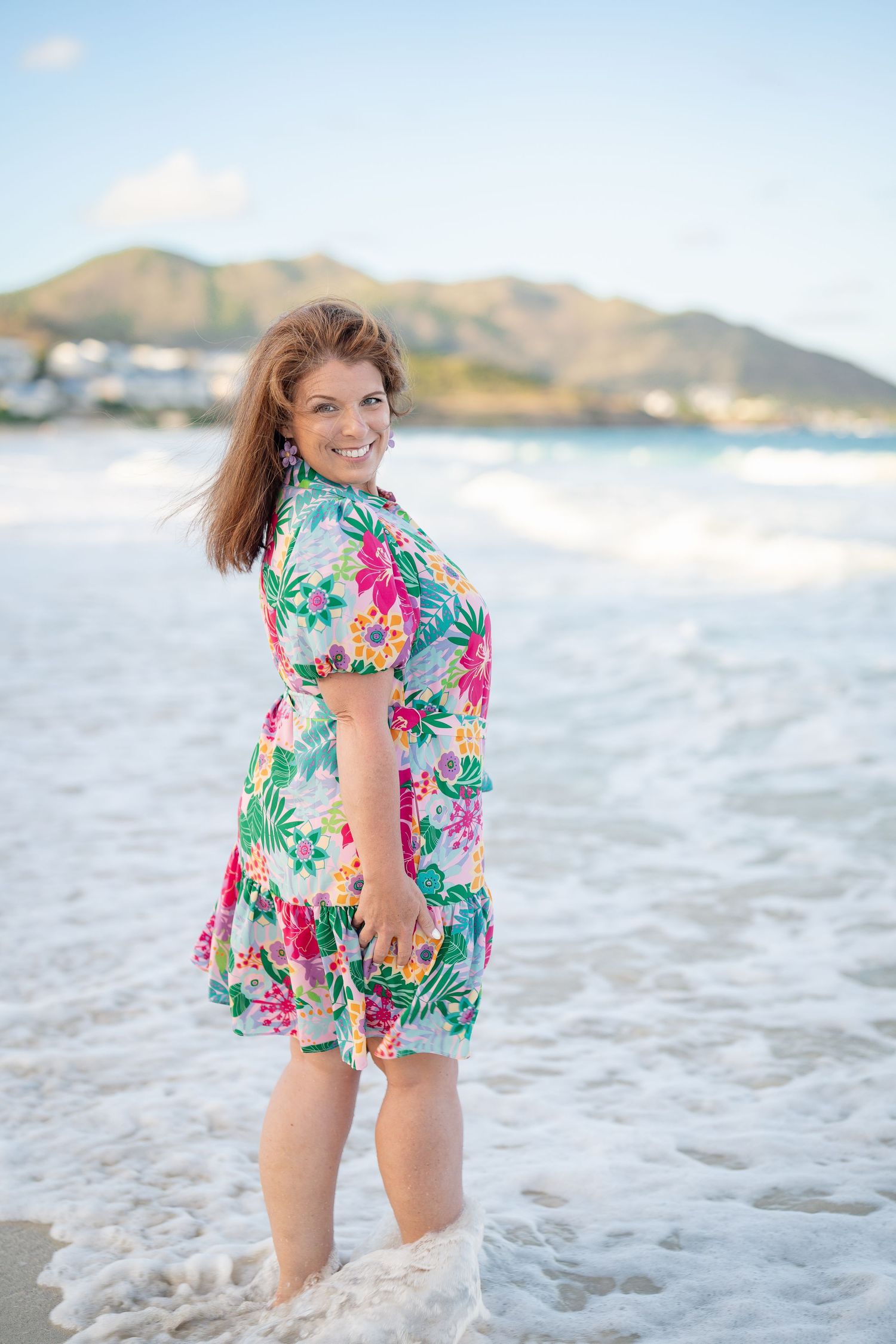 Smiling woman in colorful floral dress standing in ocean waves on sunny beach.