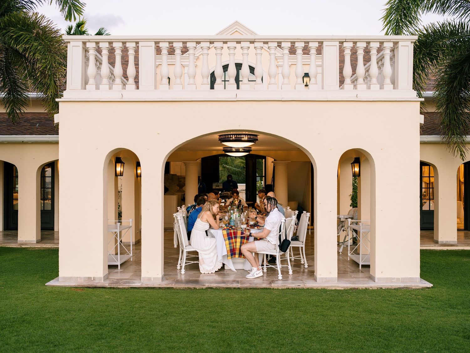 Wedding guests dining outdoors under elegant white arched villa pavilion on lush green lawn.