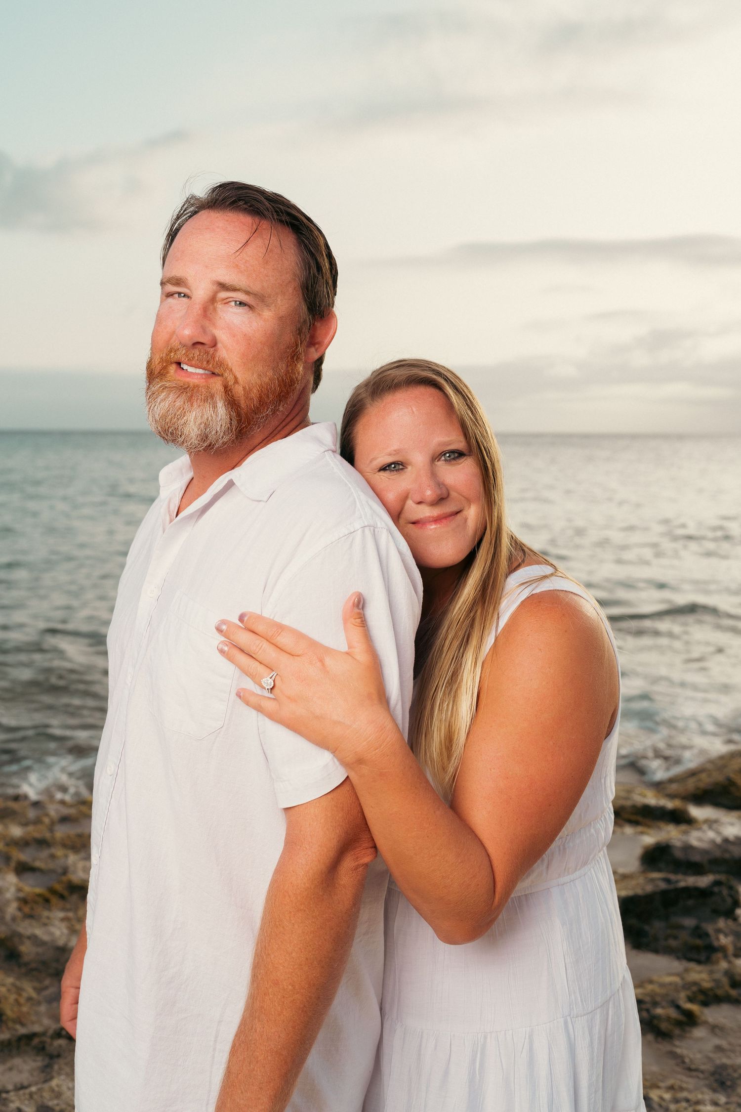 A smiling couple in white outfits embraces on rocky shoreline at sunset, woman showing engagement ring.