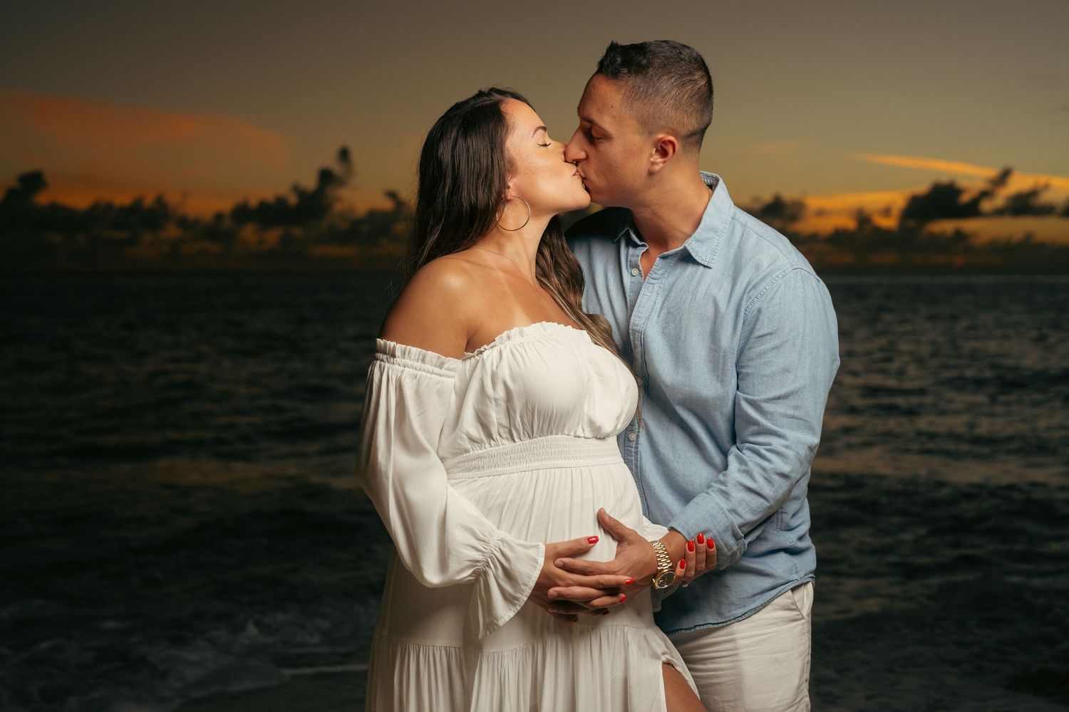 Couple kissing on beach at dramatic sunset, pregnant woman in white off-shoulder dress.