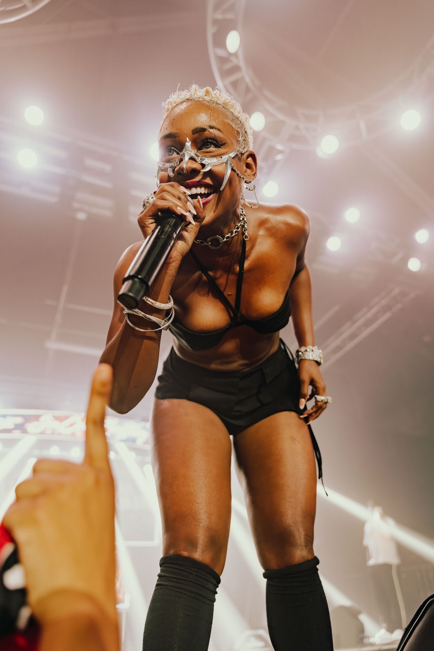 Female performer smiles while singing into microphone on stage wearing black outfit and thigh-high boots.