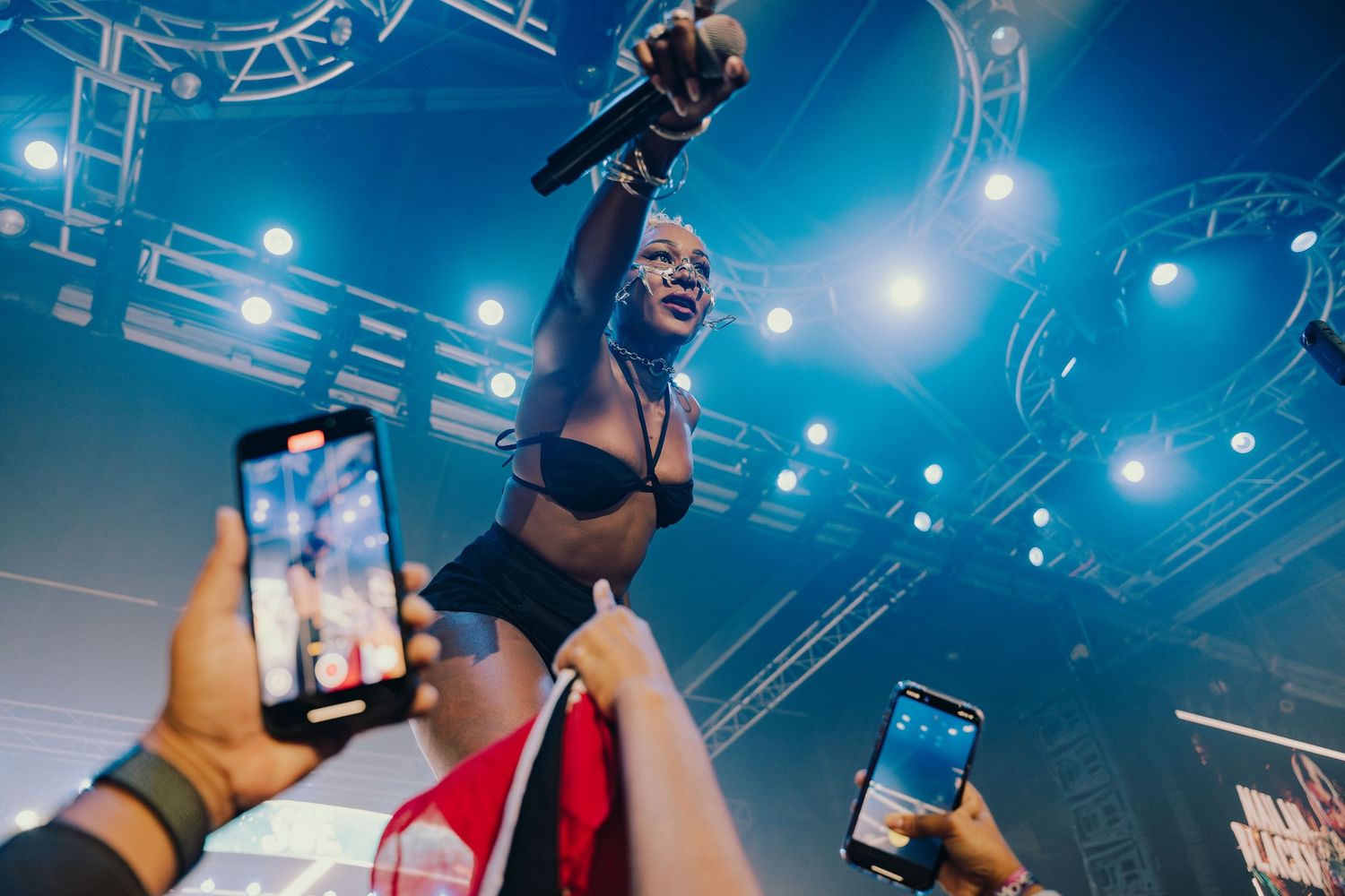 Female artiste performs on stage holding microphone while fans record with smartphones at live concert.