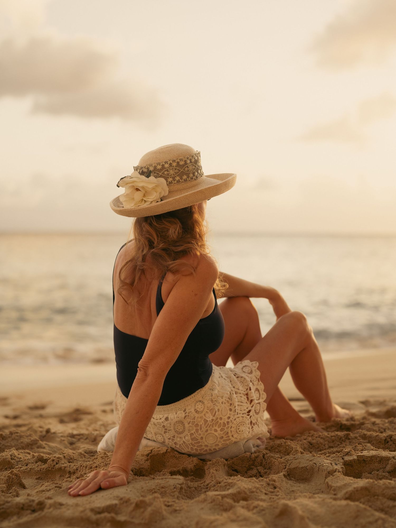 Woman in black swimsuit and decorated straw hat sitting on beach at golden sunset.