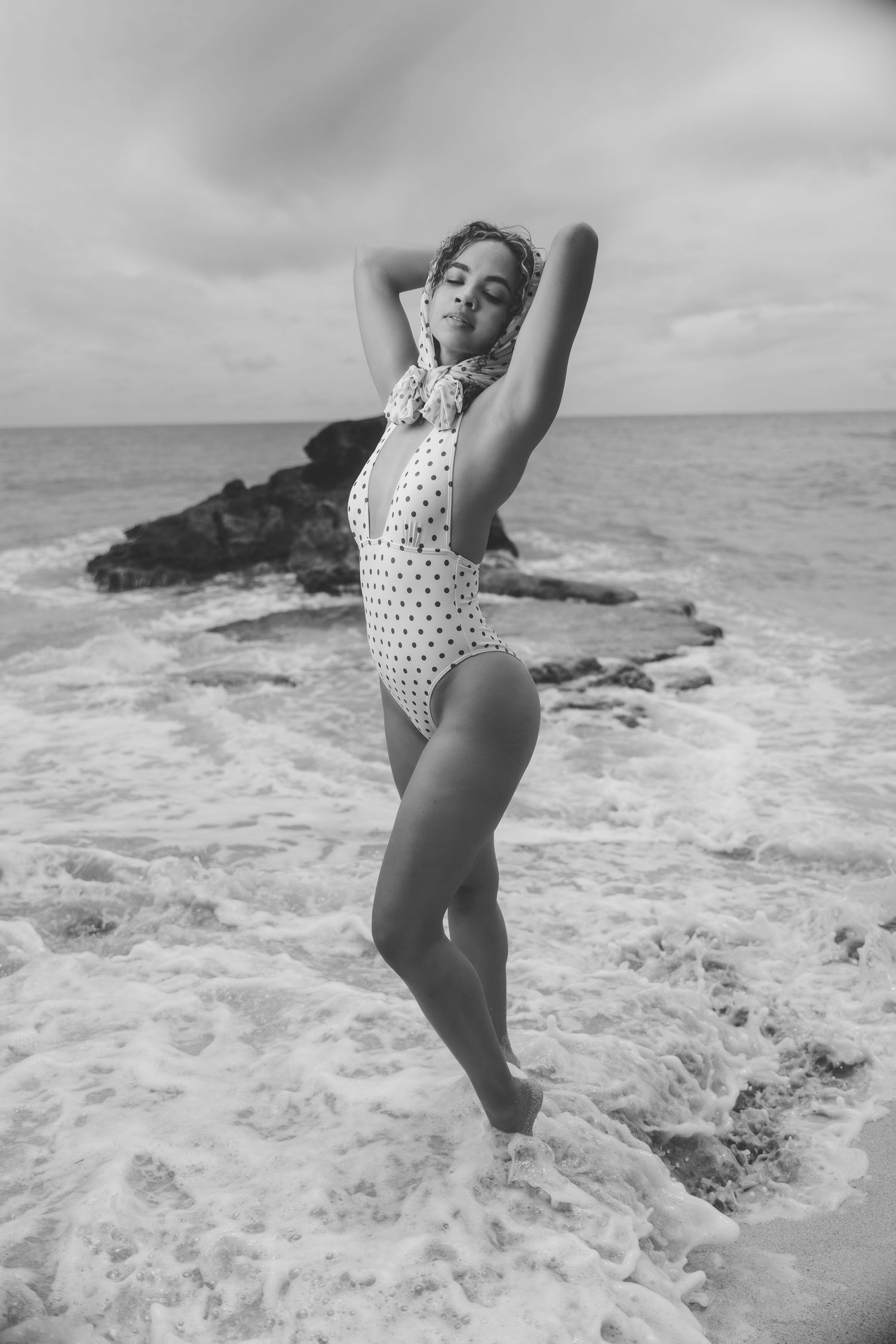 Black and white photo of woman in polka dot swimsuit posing in ocean waves near rocky shore.